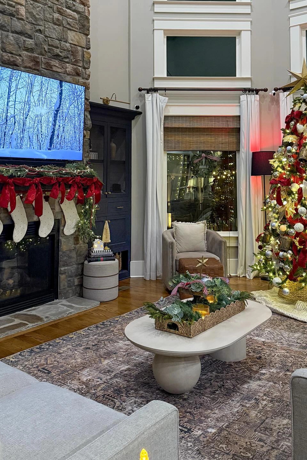 christmas living room with stone fireplace decorated with greenery garland and white stockings hanging from red ribbons, light gray armchairs around oval white coffee table with green centerpiece, beige patterned rug, tall windows with woven blinds and white curtains, christmas tree with red and gold ornaments