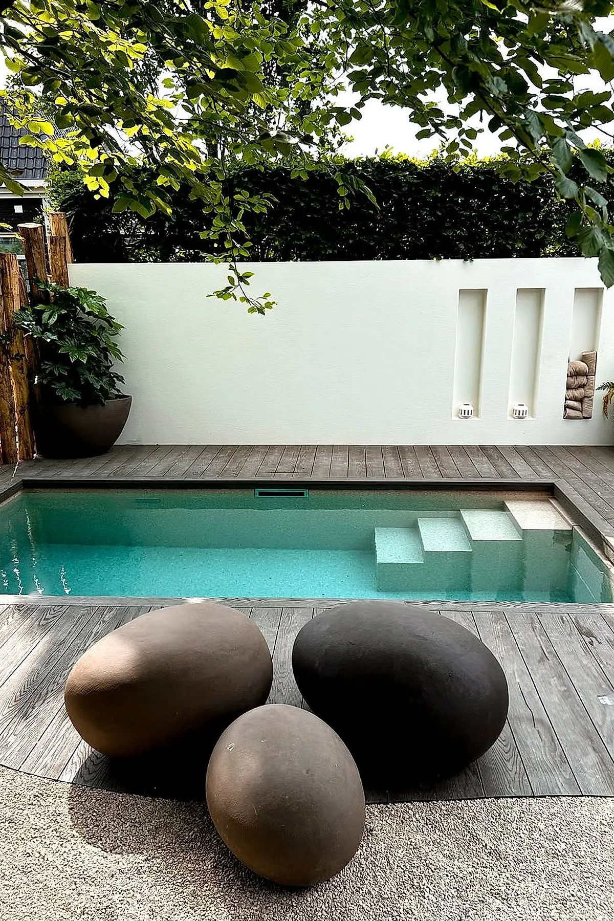 A small rectangular inground pool with clear blue water, surrounded by wooden decking and a white wall with built-in niches; in front are three large smooth stone-shaped seats in shades of brown and black, with green leafy branches overhead.