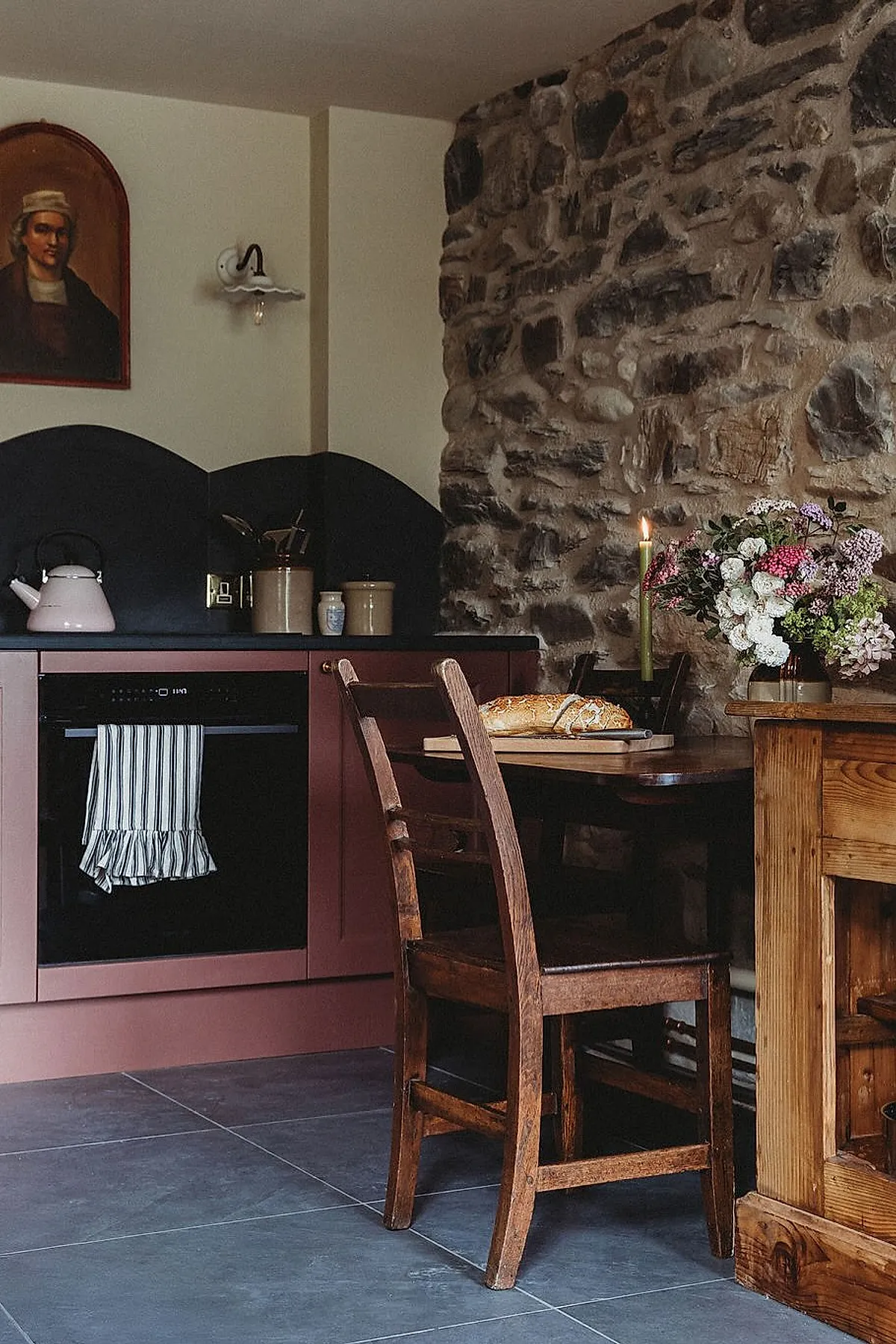 Country cottage kitchen featuring a rustic stone wall, mauve cabinets with black countertop and oven, wooden dining table with loaf of bread on cutting board, bouquet of flowers in glass vase, wooden chairs, and soft lighting from a wall sconce.