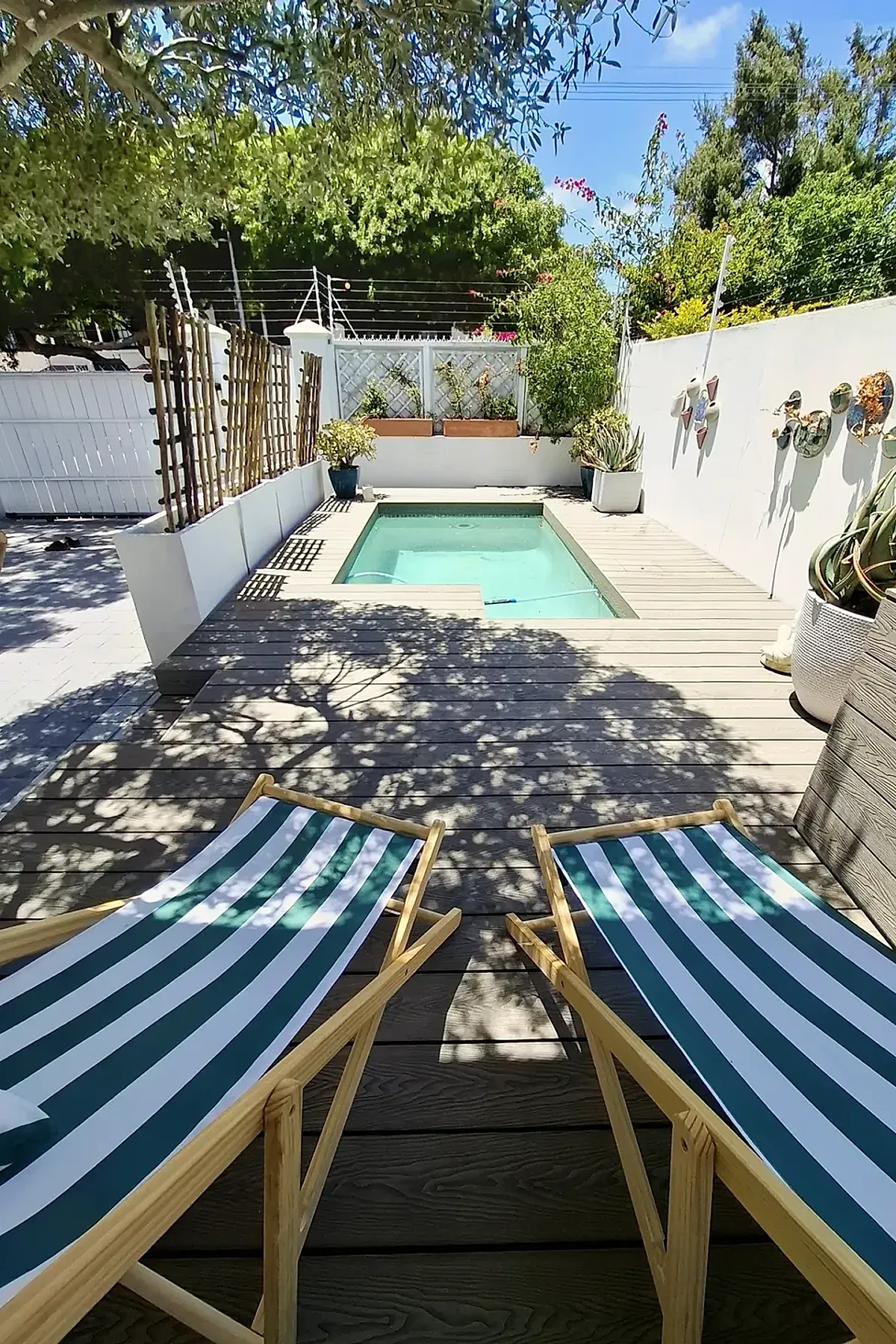 Small rectangular swimming pool surrounded by a light wood pool deck featuring two wooden lounge chairs with green and white striped fabric, potted plants along white walls, and lattice fencing under a sunny blue sky.