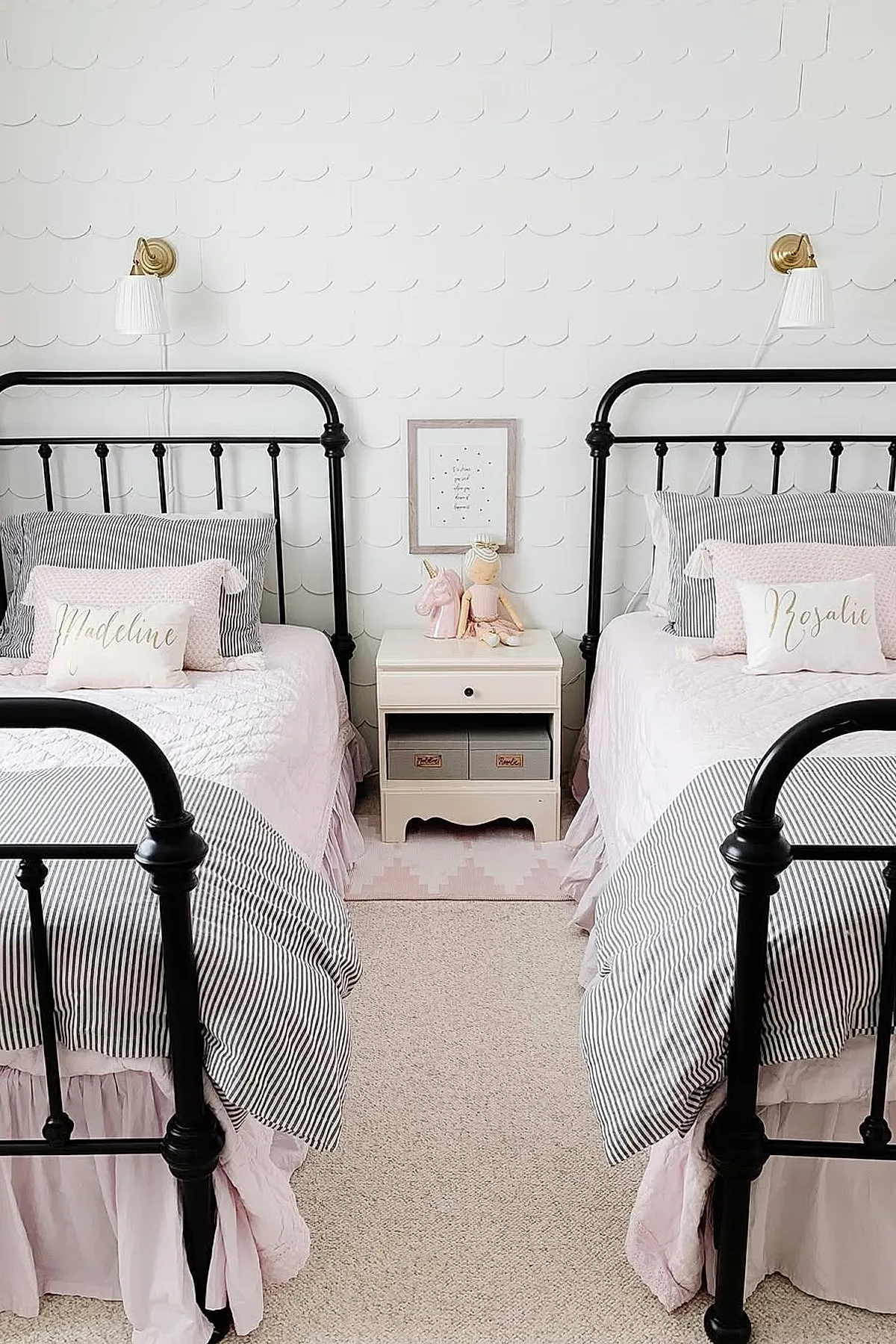Girls shared bedroom with two black metal frame beds dressed in soft pink bedding and striped pillowcases, separated by a white nightstand holding pink unicorn and doll figurines, against a white textured wall with gold sconces above each bed.