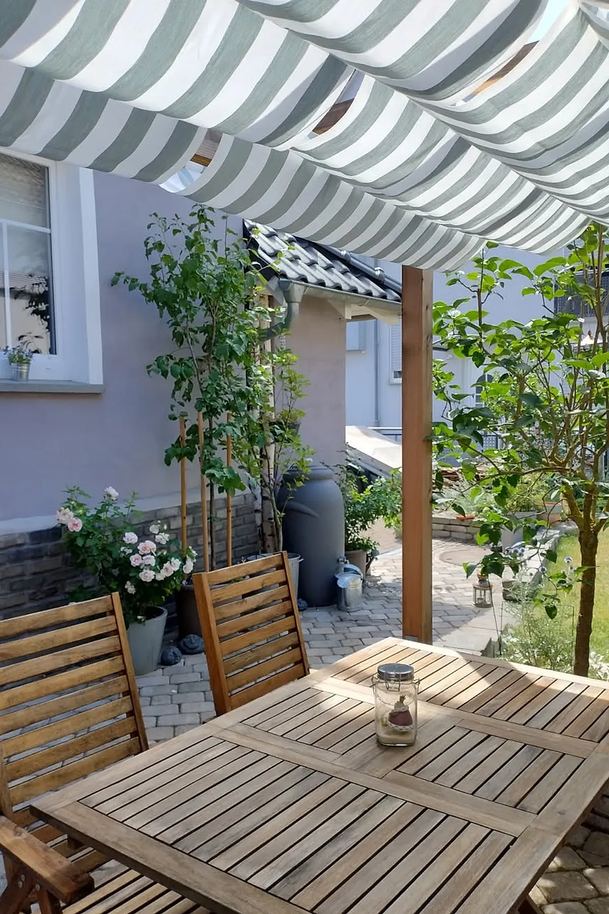 Small outdoor patio with a wooden dining table and chairs beneath a green and white striped fabric canopy, surrounded by potted plants including roses, with a stone paved floor and light-colored house wall in the background.