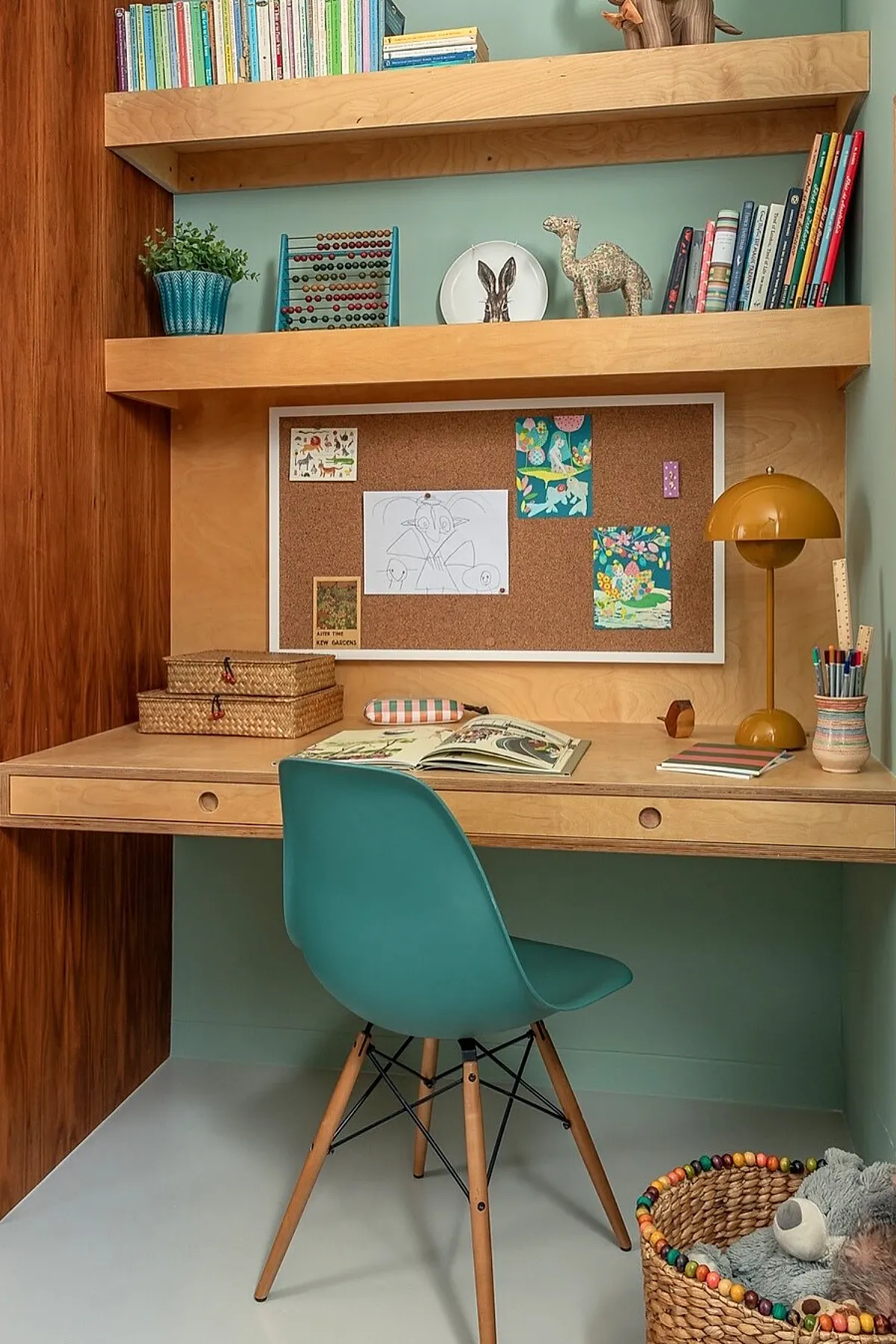 Kids study desk area in bedroom featuring a light wood floating desk with built-in drawers, teal plastic chair with wooden legs, cork bulletin board with colorful drawings pinned up, two floating shelves above holding books and decor items including an abacus and animal figurines, mustard yellow desk lamp, and woven baskets for storage.