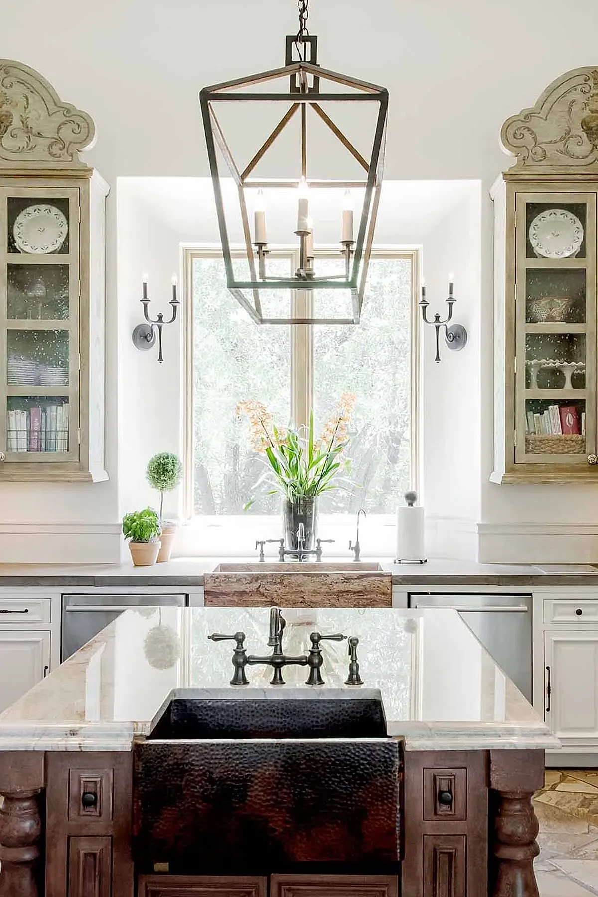 Spanish style kitchen featuring a hammered copper farmhouse sink on a dark wooden island, white cabinetry with glass doors displaying dishes and books, large window letting in natural light, and an iron lantern pendant light overhead.