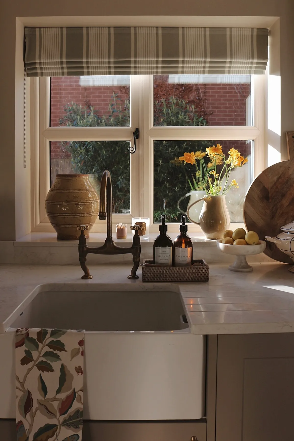 Summer kitchen decor featuring a white farmhouse sink beneath a window with beige and white striped Roman shade, rustic brown ceramic vase, cream pitcher with yellow flowers, bowl of lemons on marble countertop, and brass faucet.