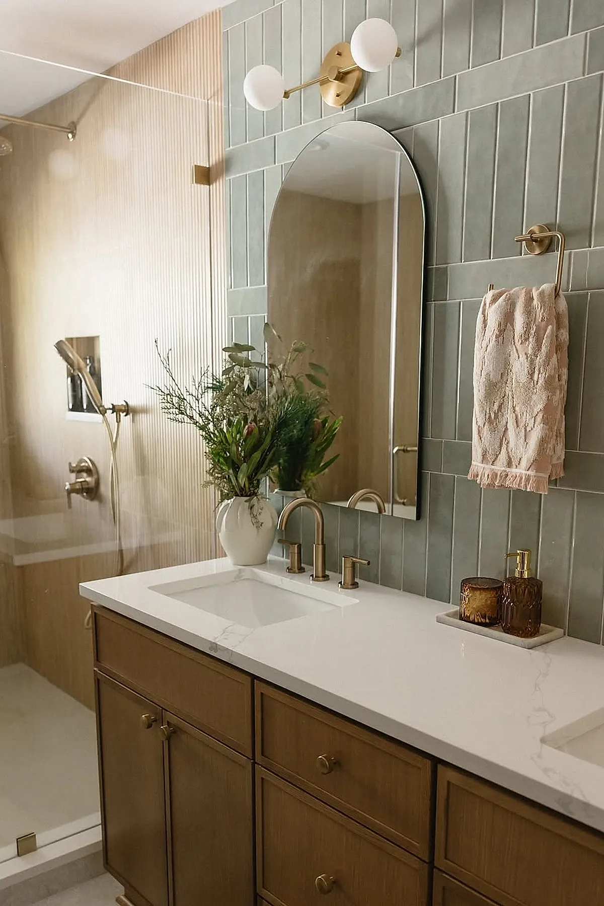 Master bathroom featuring wood cabinetry with brass knobs, white marble countertop with built-in sink, vertical muted green wall tiles behind an arched mirror, brass faucet, brass towel holder with textured towel, glass shower enclosure with brass fixtures, and a vase of fresh greenery on the counter.