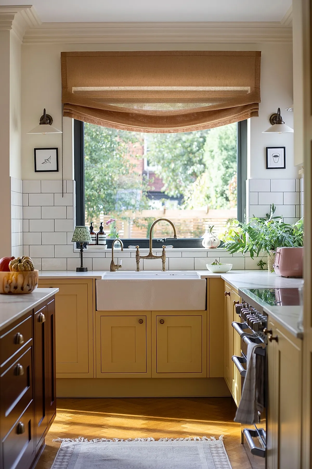 Yellow cottage kitchen featuring light yellow kitchen walls and cabinetry with brass knobs, a large white farmhouse sink under a window framed by tan Roman shades, white subway tile backsplash, vintage-style brass faucet, dark wood cabinetry on one side, and green plants on the marble countertop.