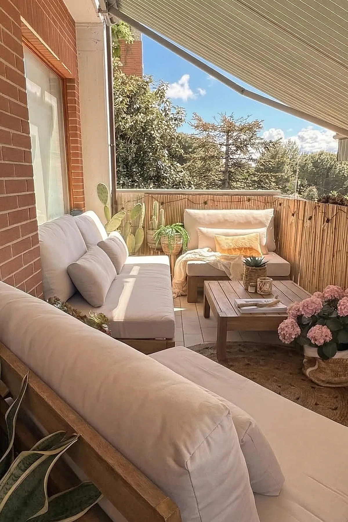 Small outdoor patio with beige cushioned wooden sofas arranged around a wooden coffee table, decorated with pink flowers in a basket, green potted plants including cacti, and a retractable striped awning overhead.