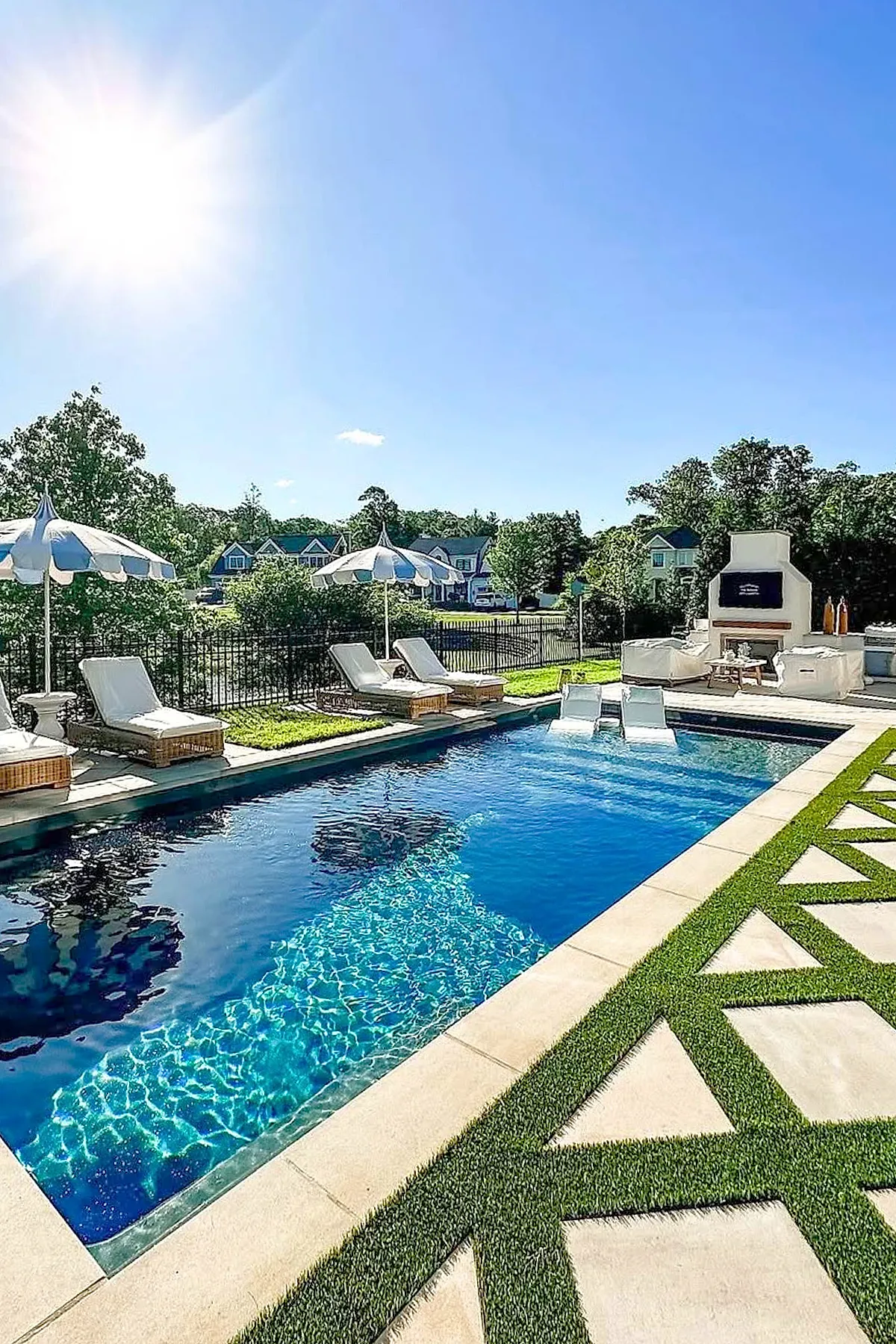 A small rectangular inground pool with sparkling blue water surrounded by a concrete deck featuring white cushioned lounge chairs and striped blue-and-white umbrellas, set against a backdrop of green grass, black fence, and houses under a clear blue sky.