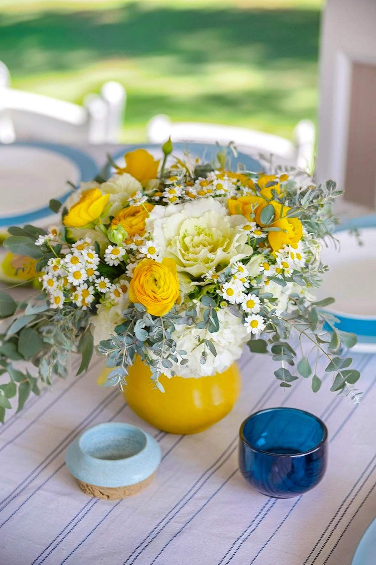 A lush, cheerful floral arrangement in a glossy, mustard-yellow ceramic vase. The bouquet includes bright yellow ranunculus, soft white ornamental cabbage, clusters of white hydrangeas, delicate feverfew flowers (small white blooms with sunny yellow centers), and silvery eucalyptus branches for texture and greenery.