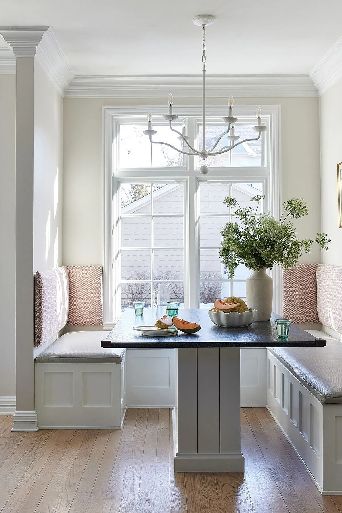 Elegant breakfast nook with pink pillows, a modern chandelier, and fresh fruit on a dark table.