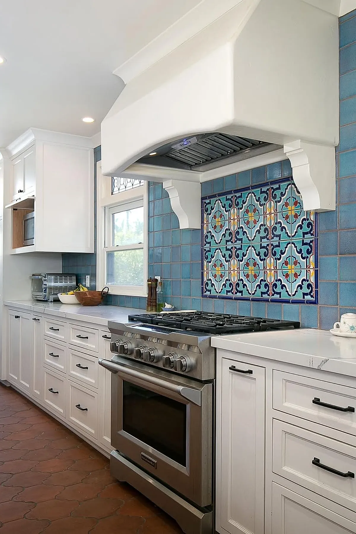 Spanish style kitchen featuring white cabinetry, a stainless steel gas stove with an ornate white range hood, and vibrant blue patterned tile backsplash framed by smaller solid blue tiles.