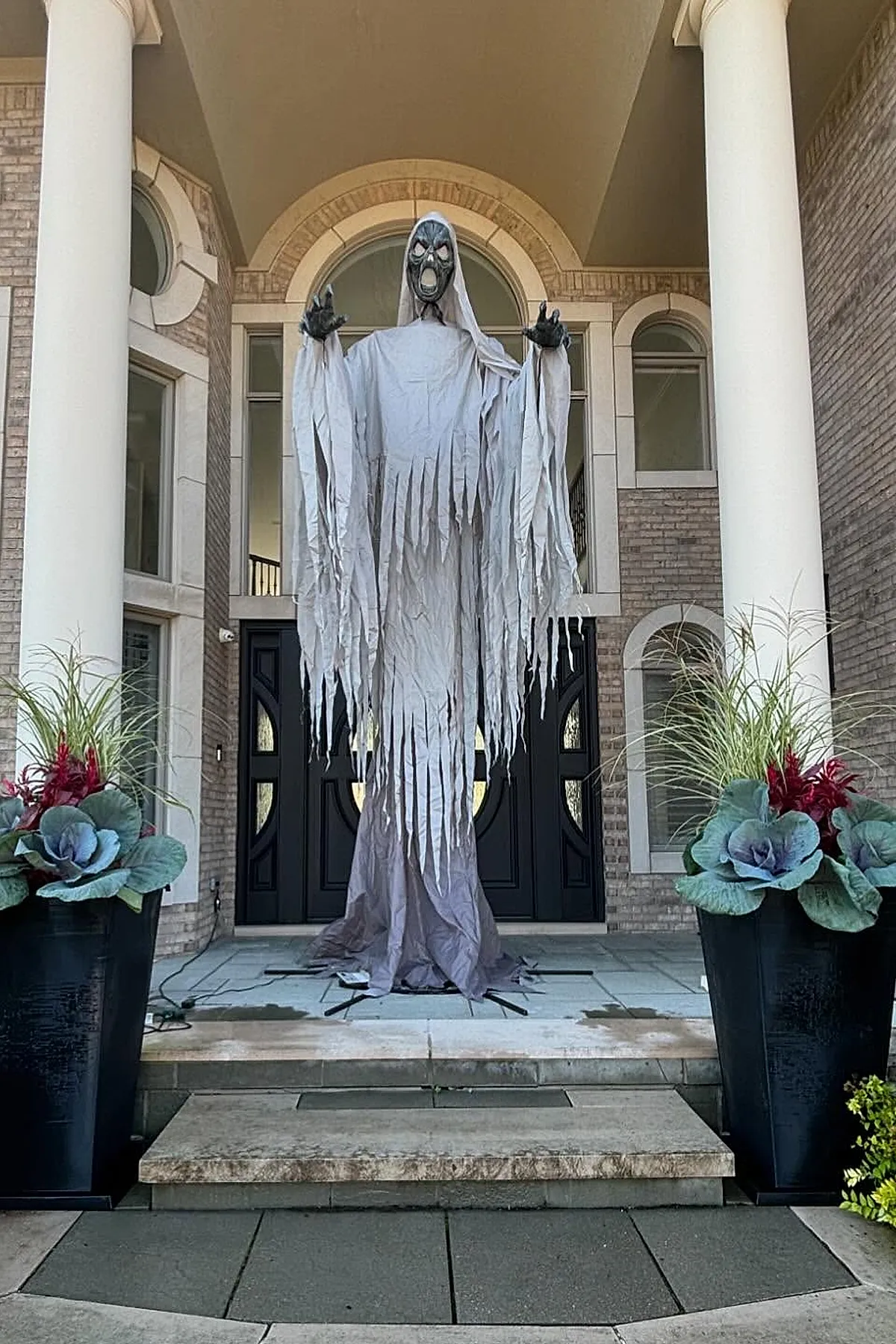 large hanging ghost decoration with torn grayish-white fabric and black hands standing on porch in front of dark double doors flanked by tall black planters with leafy green plants