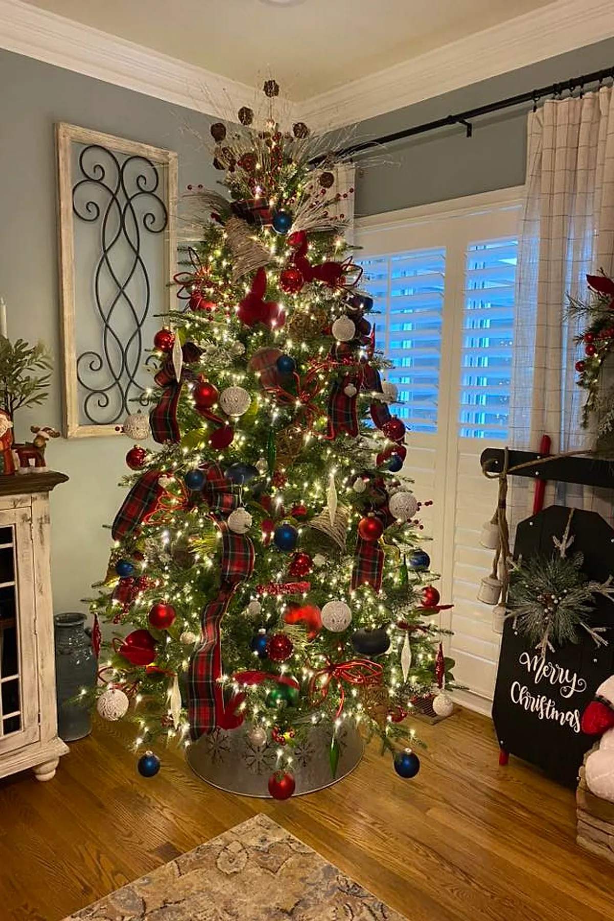 A Ralph Lauren Christmas tree decorated with red and blue baubles, white textured ornaments, plaid ribbons, and gold accents in a cozy living room with light blue walls, white shutters, wooden floor, and rustic furniture.