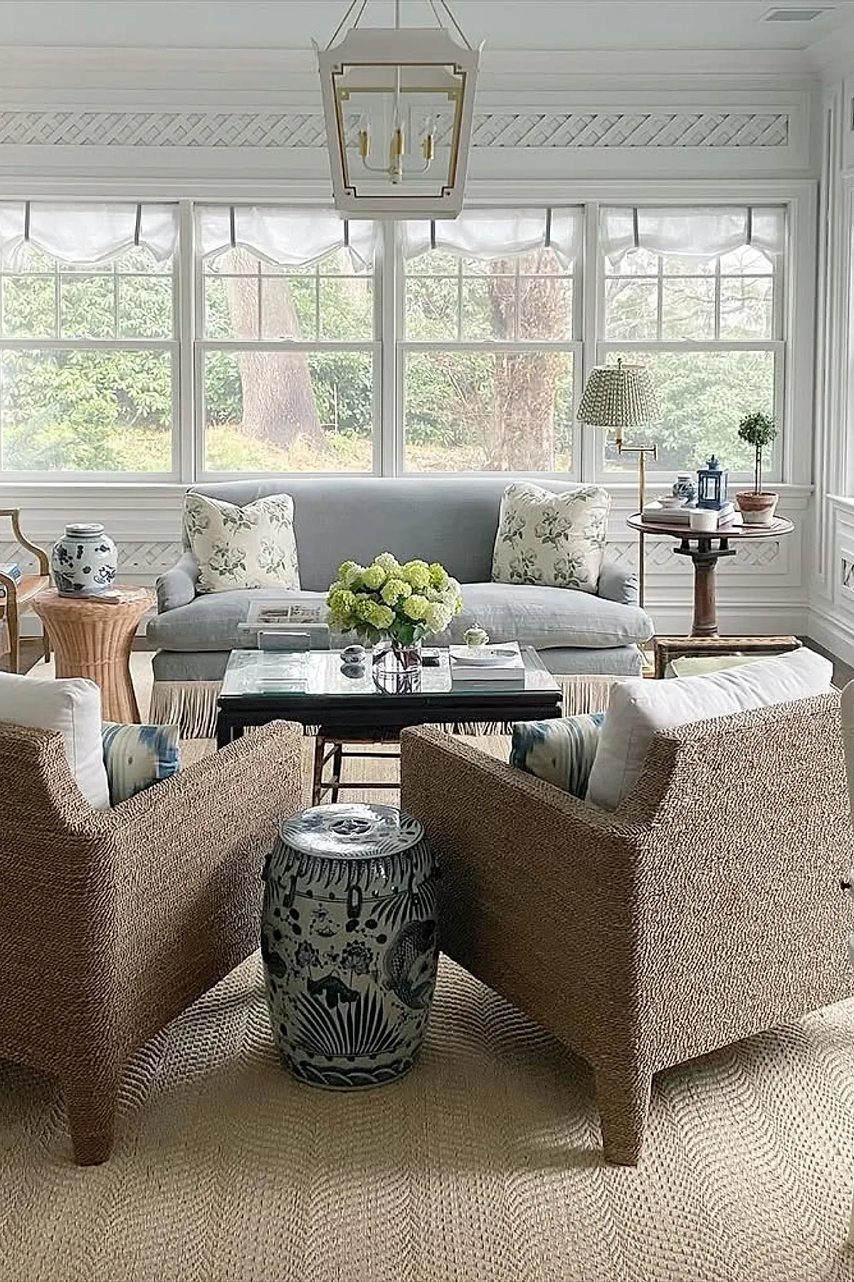Sunroom with light blue sofa, two brown rattan armchairs, white cushions, floral pillows, round side table with lamp, glass coffee table with flowers, large windows with sheer curtains letting in natural light.