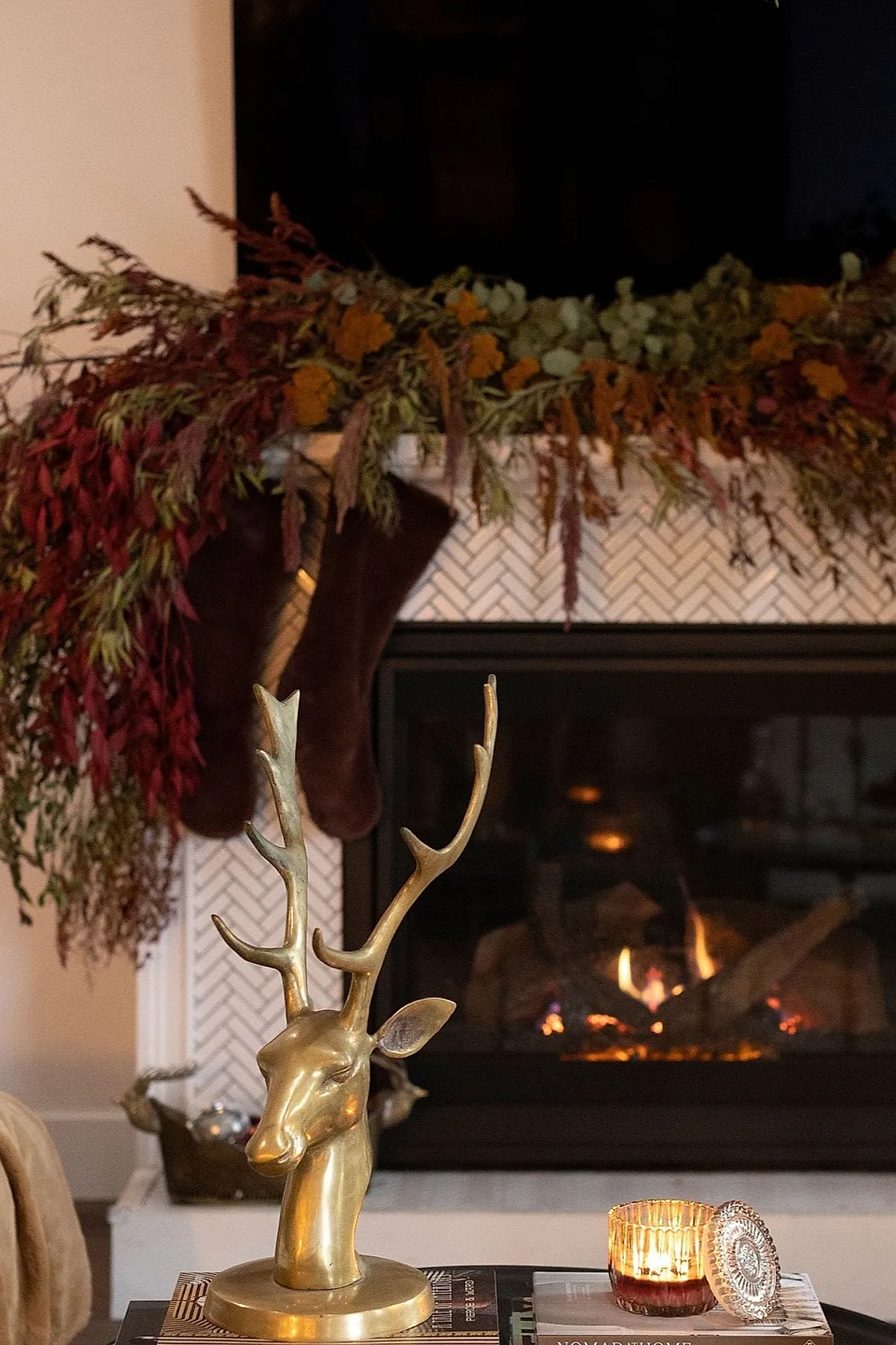A brass deer head sculpture sitting on stacked books beside a lit candle in a glass holder with a fireplace decorated with dried foliage garland and dark stockings in the background.