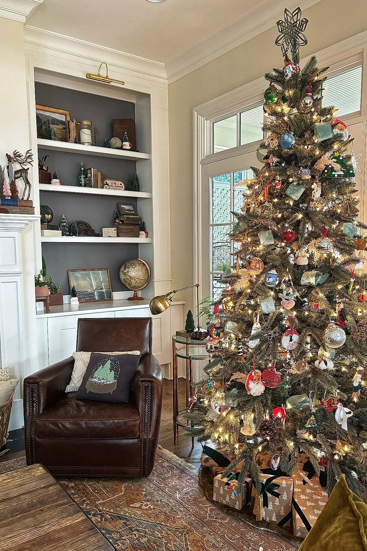 Vintage Christmas tree filled with nostalgic ornaments including Santa faces, hearts, and whimsical shapes, lit by warm white string lights beside a brown leather armchair and built-in shelves decorated with books and holiday accents.