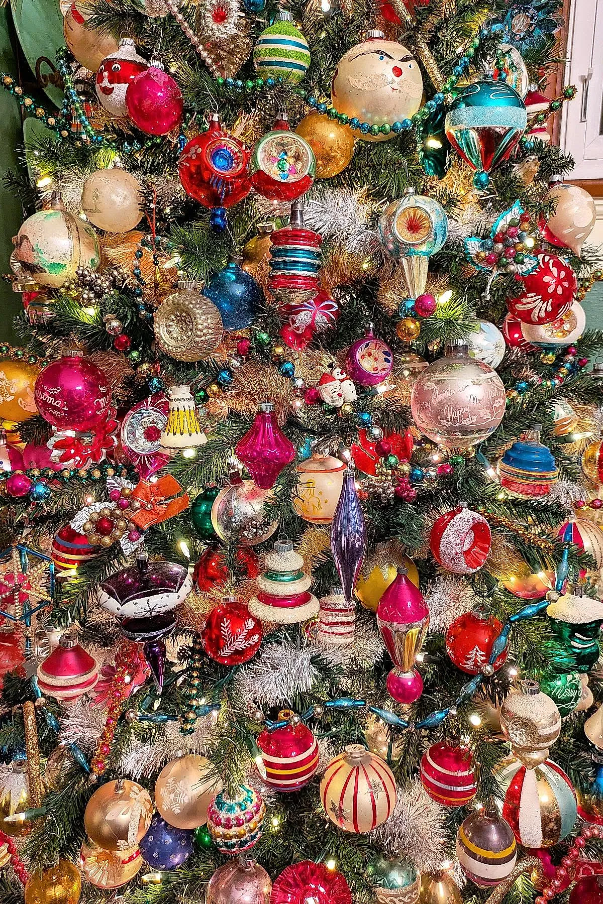 Densely decorated vintage Christmas tree with bright colorful glass ornaments in round, teardrop, and novelty shapes, surrounded by lush green pine branches and warm white lights.