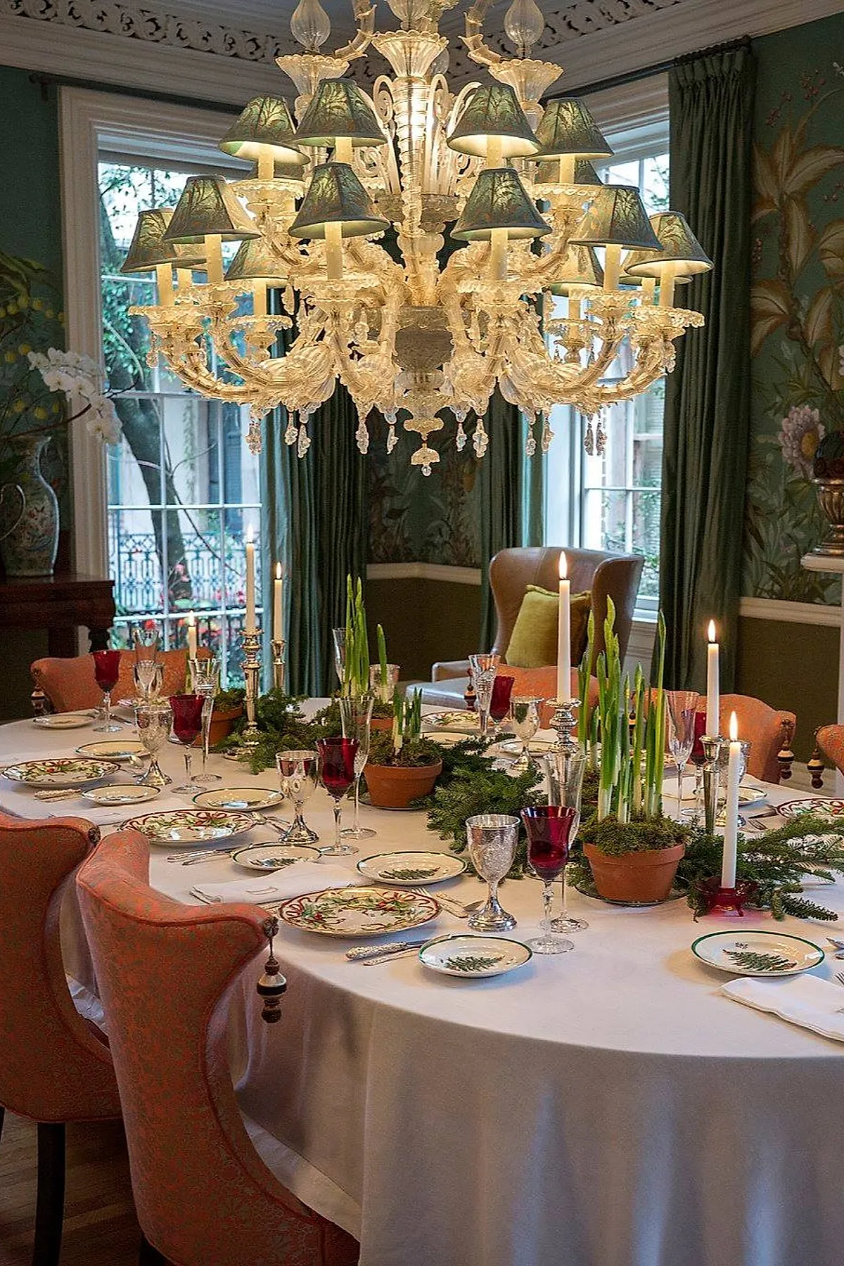 An elegant Ralph Lauren Christmas dining room featuring a round table with a white cloth, potted greenery, candles, festive plates, red glassware, coral chairs, and a crystal chandelier.