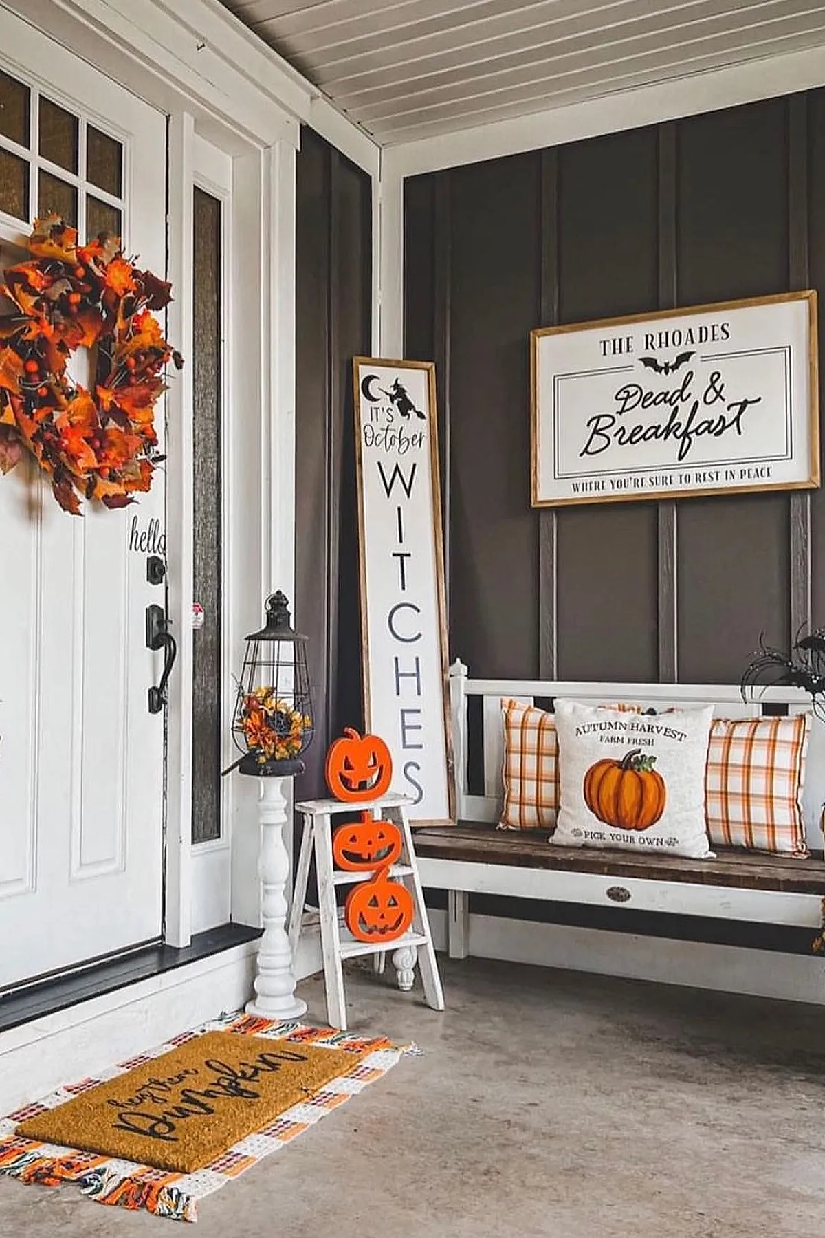 three orange carved pumpkins stacked on white wooden ladder next to black lantern with fall flowers white bench with plaid and pumpkin pillows tall Halloween sign and autumn leaf wreath on white door