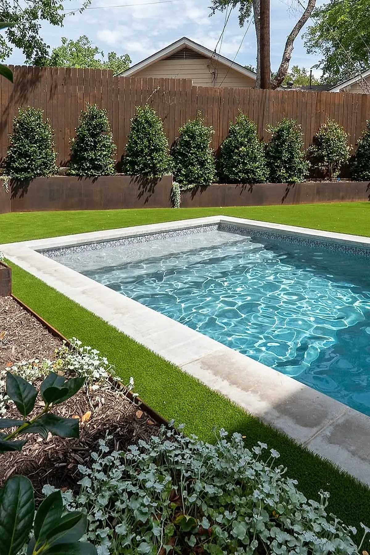 Rectangular plunge pool with clear blue water surrounded by white stone edging and bright green artificial turf, backed by a rust-colored metal planter box filled with evenly spaced green shrubs, all enclosed by a wooden fence under a partly cloudy sky.