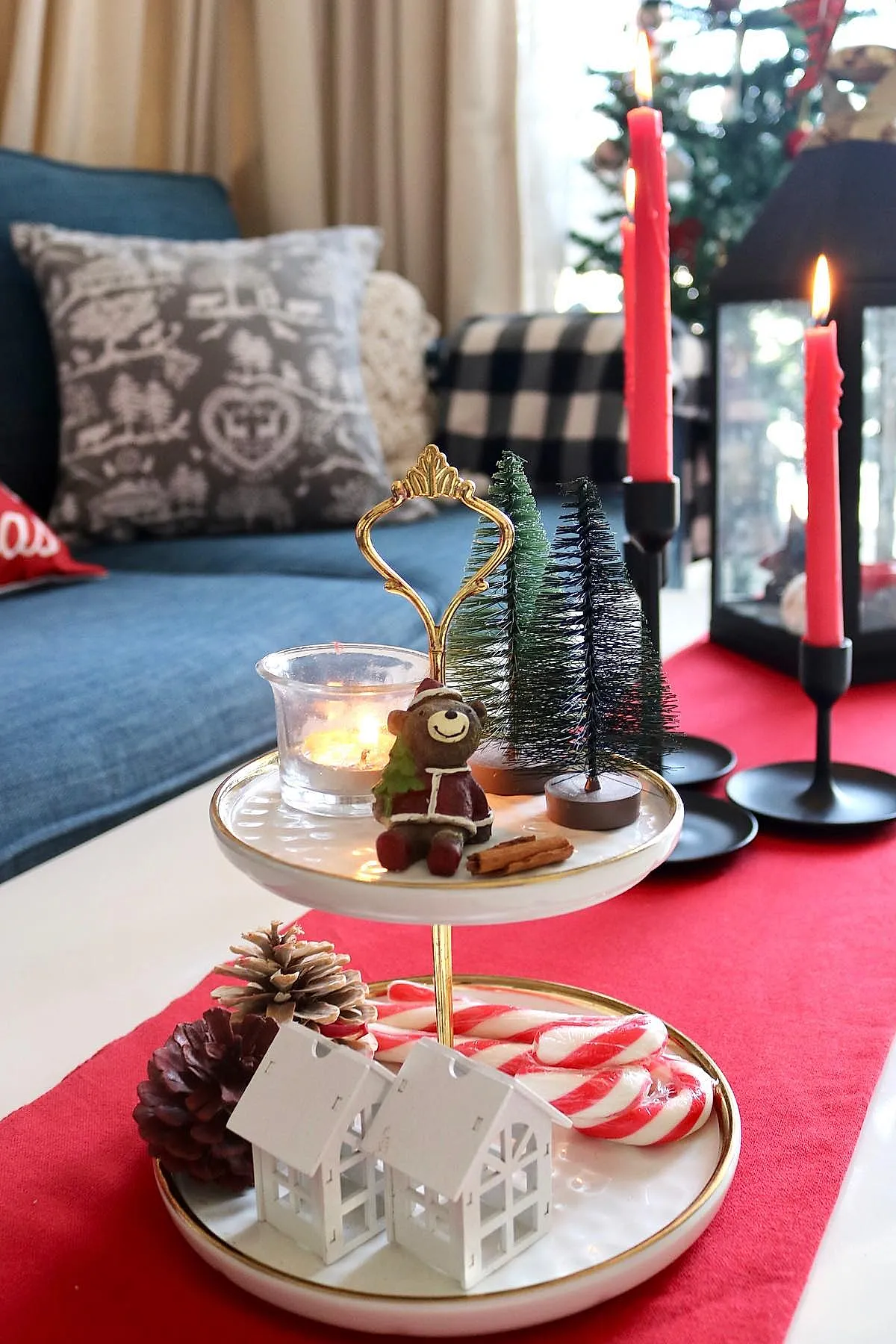 two-tiered ceramic tray with gold handle holding miniature bottle brush Christmas trees, a small smiling bear figurine dressed as Santa, cinnamon sticks, pinecones, white miniature house decorations, and wrapped red and white candy canes on a coffee table with a red runner, set against a blue sofa and black candle holders with red candles.
