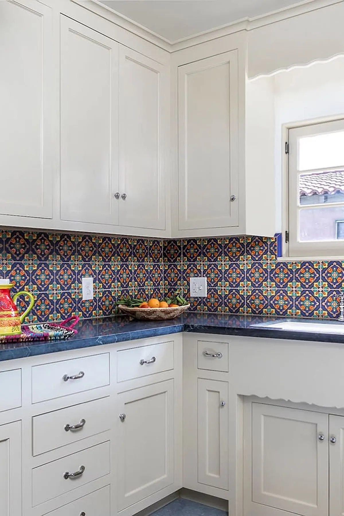 Spanish style kitchen with white cabinetry, colorful patterned tile backsplash featuring navy blue, orange, and teal, dark countertop, silver cabinet handles, and a window with a scalloped valance showing roof tiles outside.