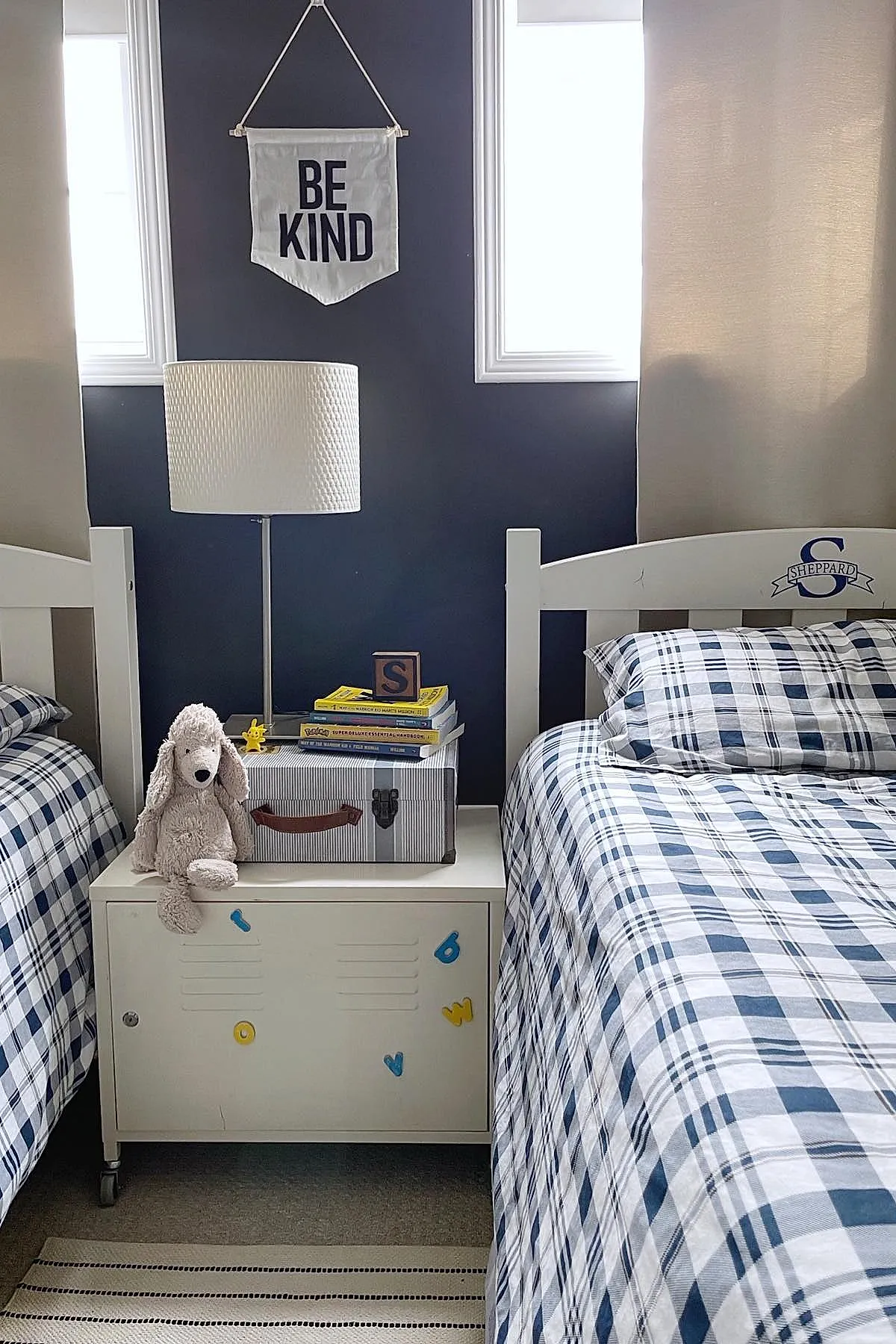 Two twin beds with navy blue and white plaid bedding, white wooden headboards, small white nightstand between beds holding a lamp, books, a toy block, and a stuffed dog plush against a dark blue accent wall with