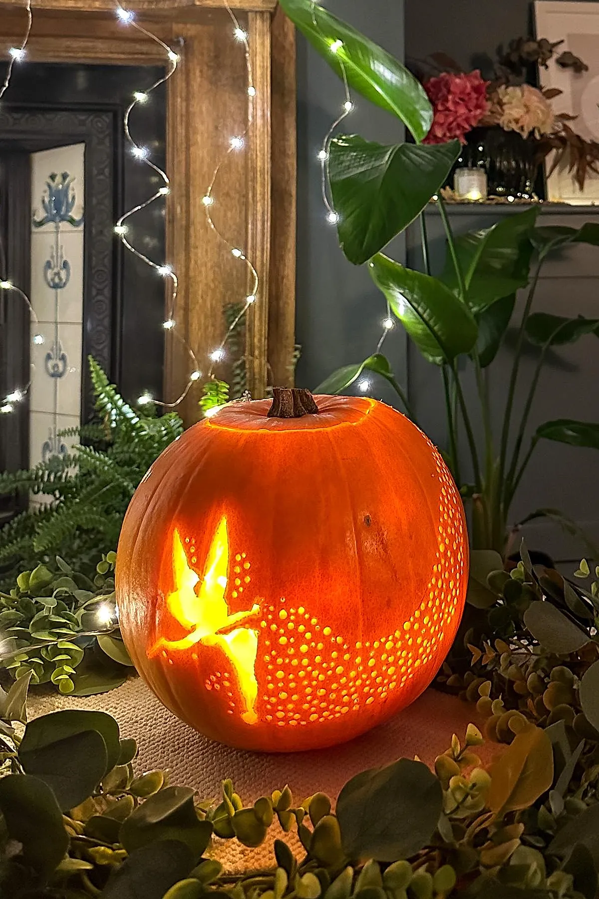 Orange pumpkin carved with glowing silhouette of Tinkerbell and surrounding trail made of small holes, set among green leaves and fairy string lights indoors.