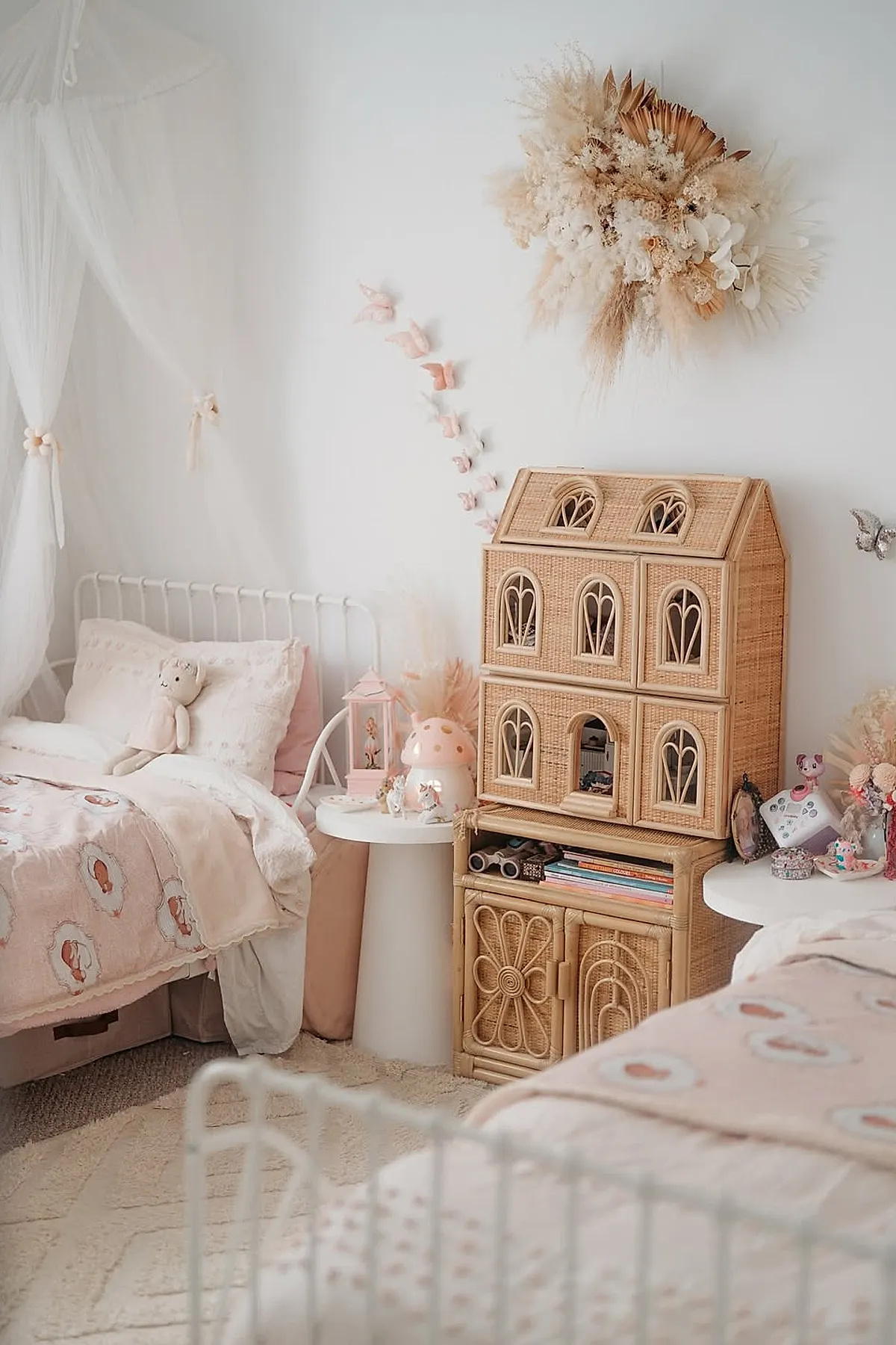 Girls shared bedroom with white metal beds, soft pink bedding, a large rattan dollhouse on a matching rattan cabinet, white side tables, and delicate butterfly wall decor on white walls.