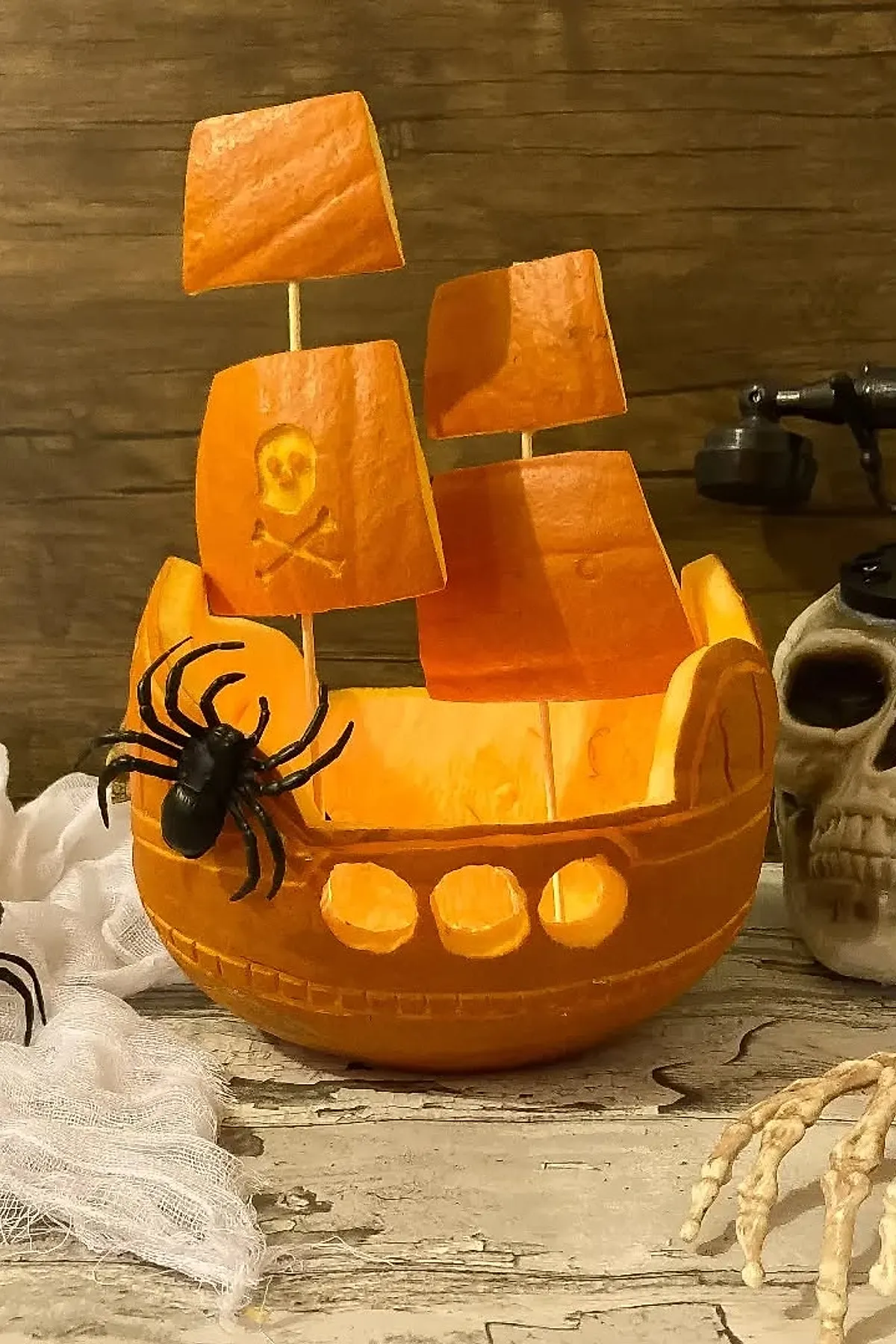 Orange pumpkin carved as a pirate ship with three rectangular sails, one showing a skull and crossbones, decorated with black plastic spider, next to a skull decoration and skeleton hand on weathered wood.