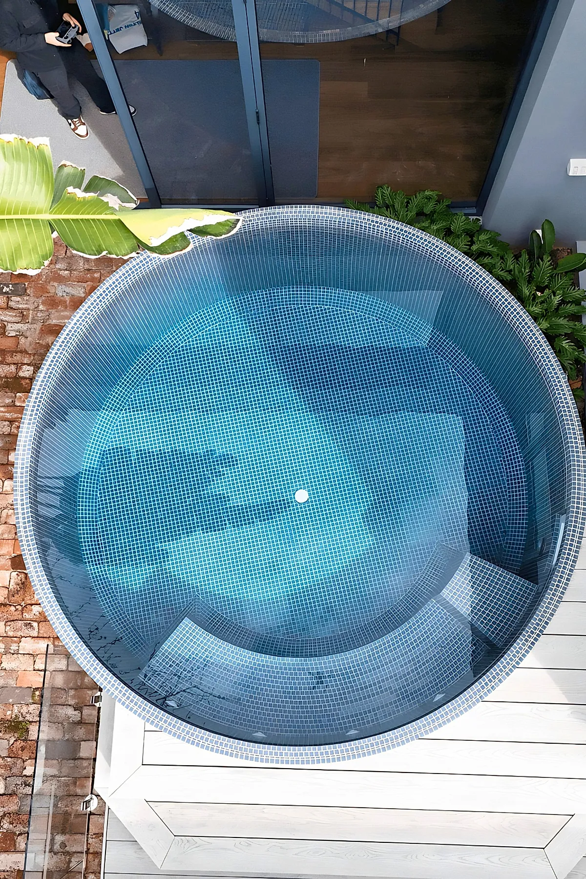 Round above ground plunge pool with blue mosaic tiles, built into a white wooden deck, surrounded by brick paving and lush green plants near a glass sliding door.