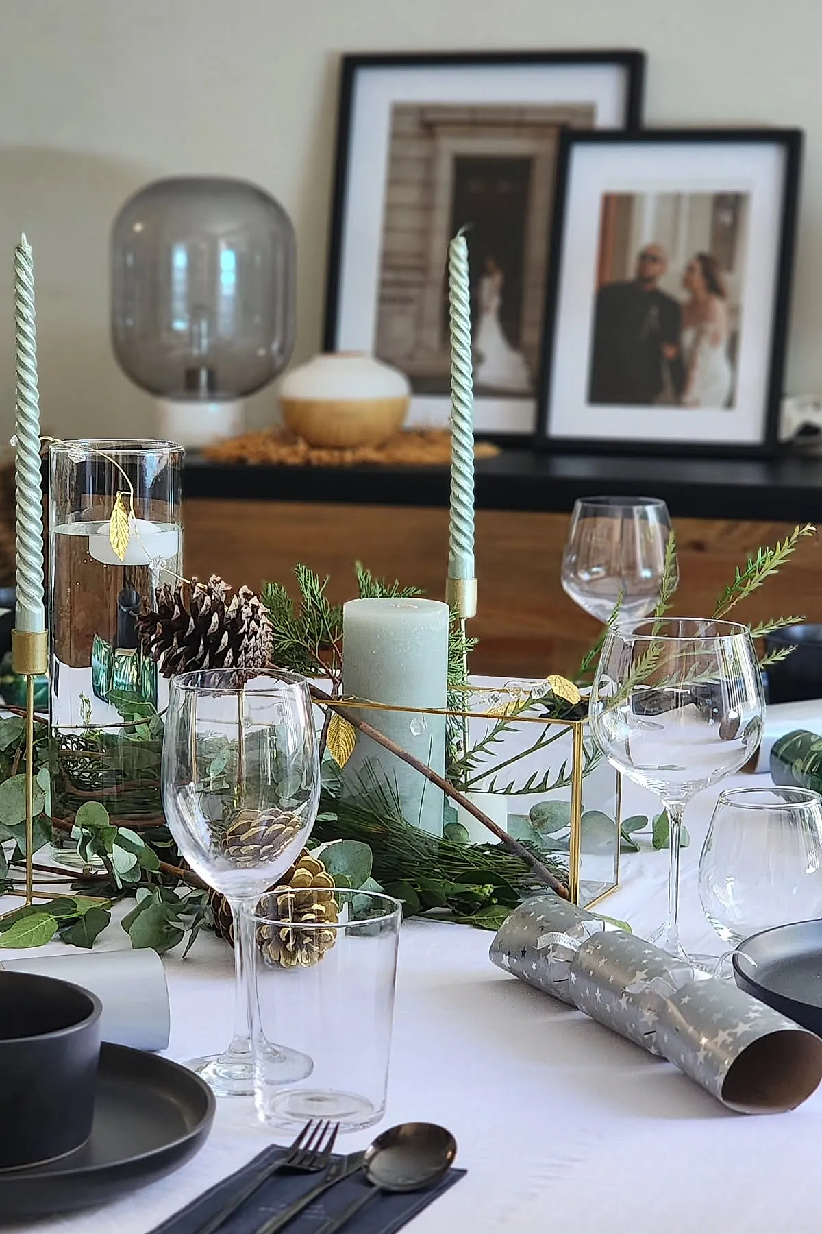christmas dinner table settings with twisted green candles, greenery garland, pinecones, clear wine glasses, silver Christmas crackers, black matte plates, and silver cutlery on a white tablecloth.