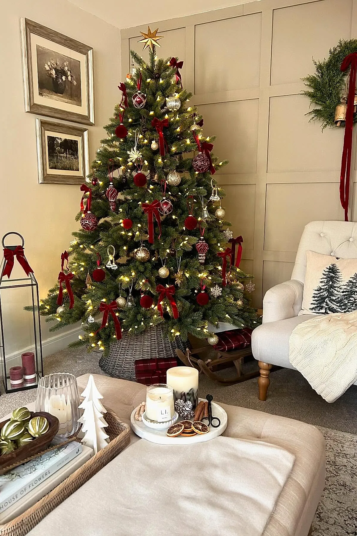 A traditional christmas tree decorated with red velvet bows, gold glitter balls, various glass ornaments including patterned globes and snowflakes, all illuminated by warm white string lights in a cozy living room.