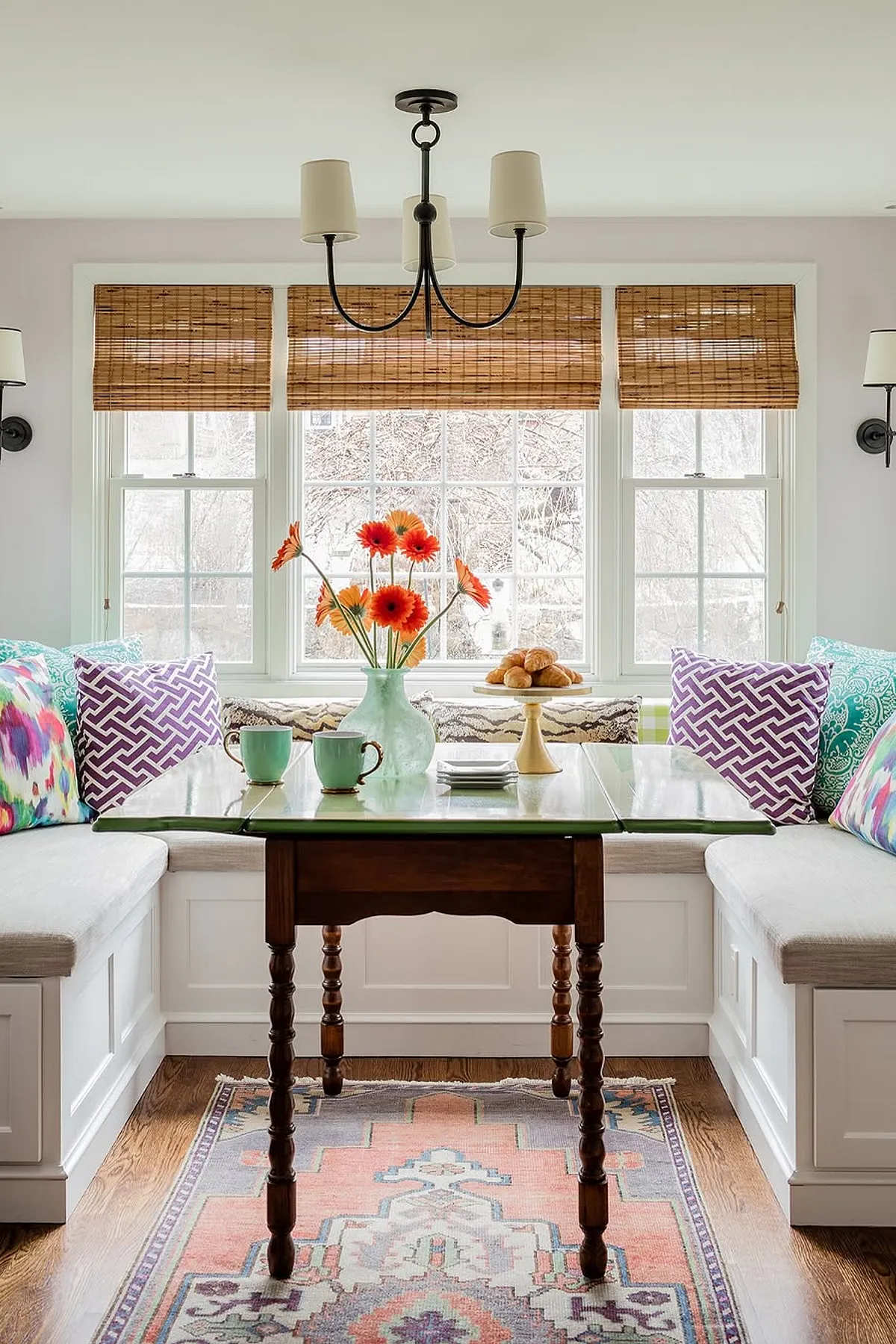 Bright breakfast nook featuring colorful cushions, a wooden table, fresh flowers, and natural light.