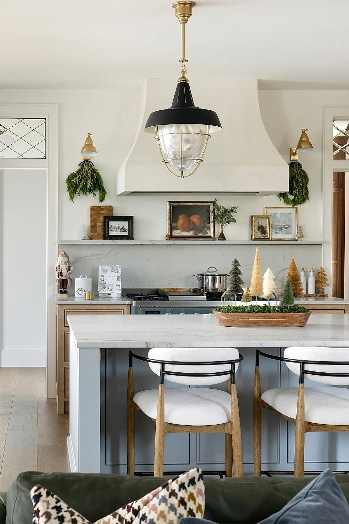 neutral minimalist christmas decor featuring a wooden tray filled with various sized bottle brush trees in white, green, and gold tones placed on a white marble kitchen island with light blue cabinetry and wood floor
