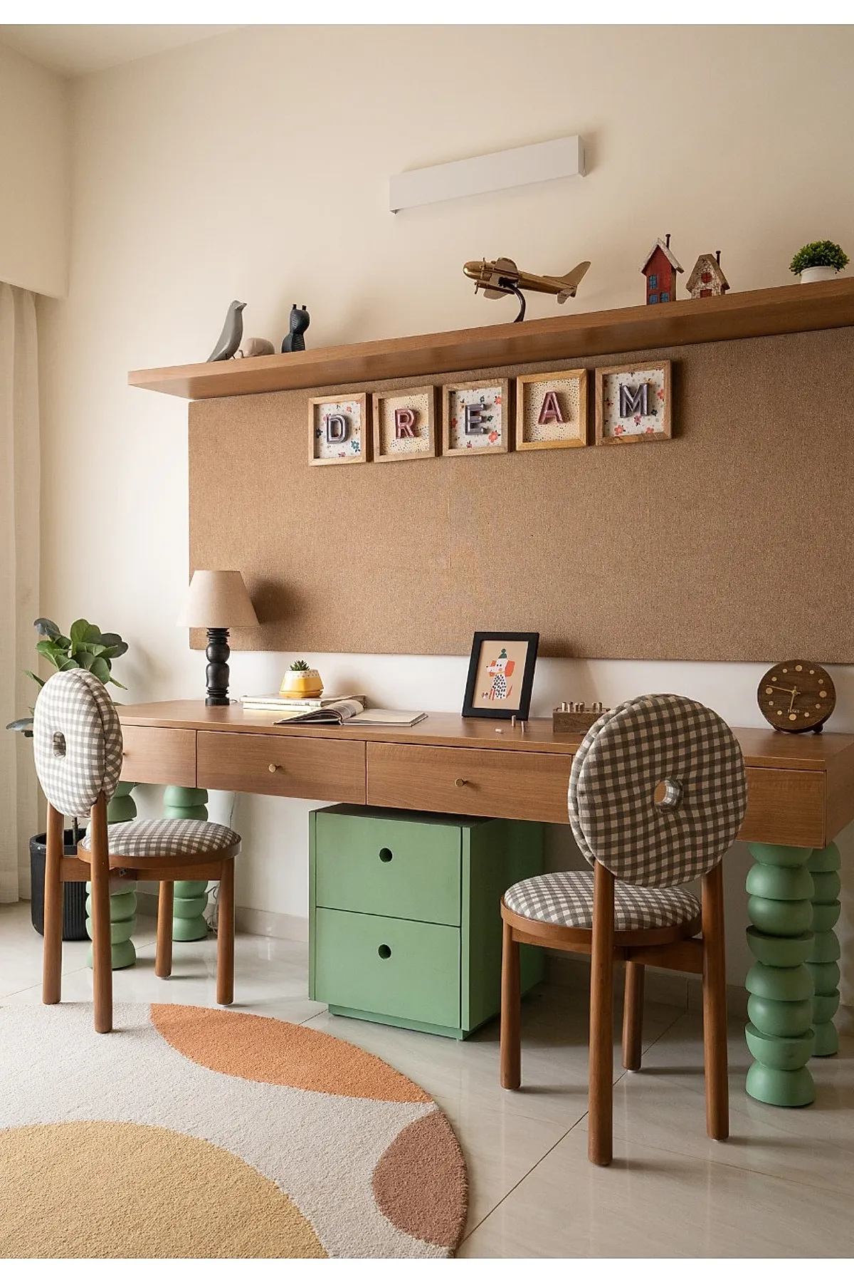 Kids desk area in bedroom featuring a wooden study desk with chunky green bubble-style legs, two round-backed chairs with brown and white gingham upholstery, a cork bulletin board with framed letters spelling
