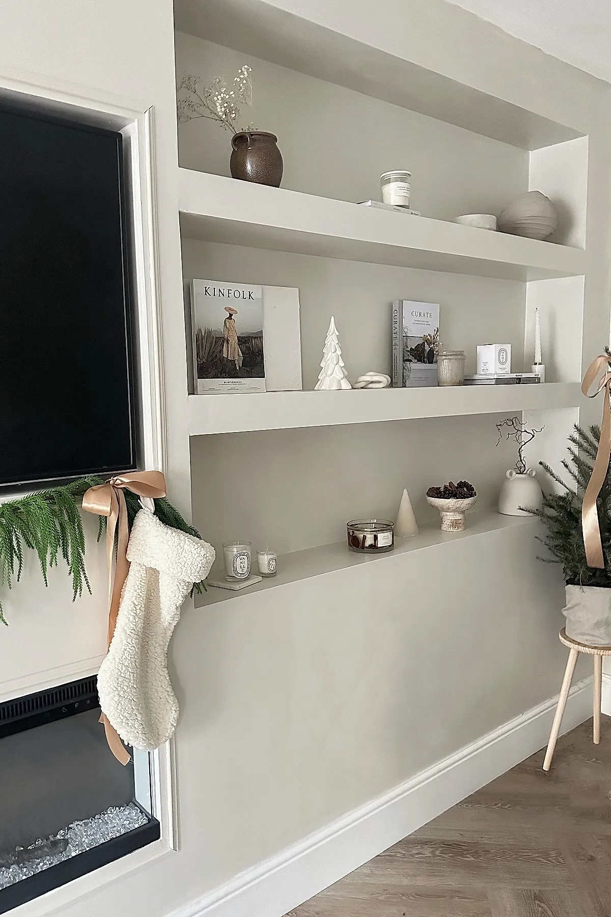 built-in neutral-toned shelves decorated with minimalist christmas decor including white ceramic trees, candles, pine cones in a pedestal bowl, books, dried flowers in vases, and a cozy cream stocking hanging beside green garland with peach satin ribbons