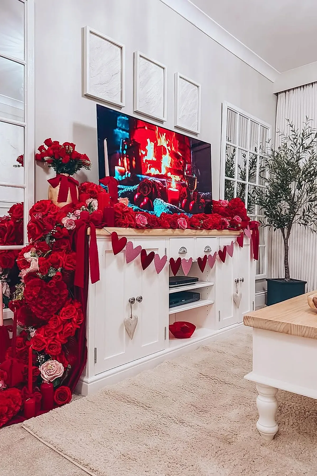 White TV stand decorated with abundant red and pink roses, red candles, and a heart garland hanging across the front. A flat-screen TV shows a cozy fire scene, complemented by neutral carpet and light-colored walls.