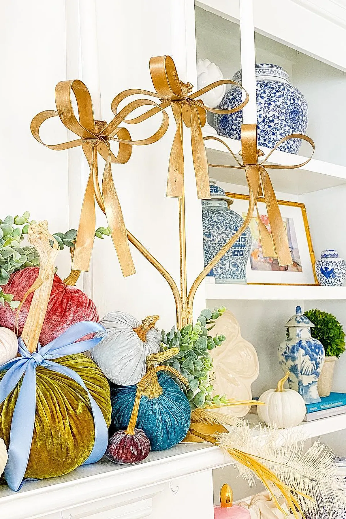 fall mantel decorated with colorful velvet pumpkins in mustard, teal, burgundy, and white shades accented with blue satin ribbons and gold metal candle holders with ribbon bows