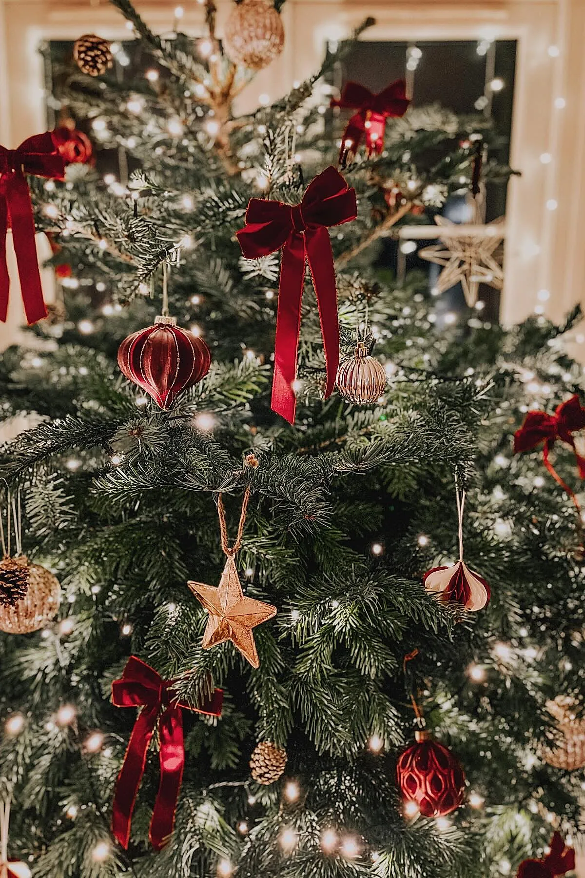 Christmas tree decorated with red velvet bows, gold star ornaments, ribbed red and gold baubles, pinecones, and warm white string lights on green pine branches