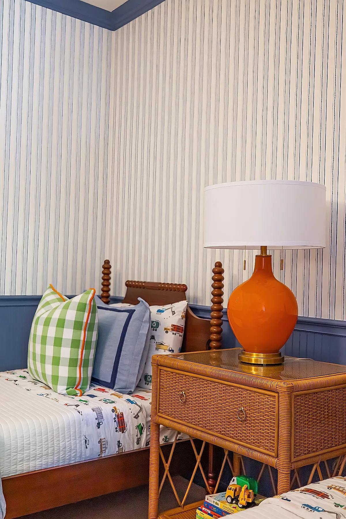 Boys bedroom with vertical blue and white striped wallpaper, wooden spindle bedframe, green check pillow, transportation print bedding, woven rattan nightstand with bright orange ceramic lamp.