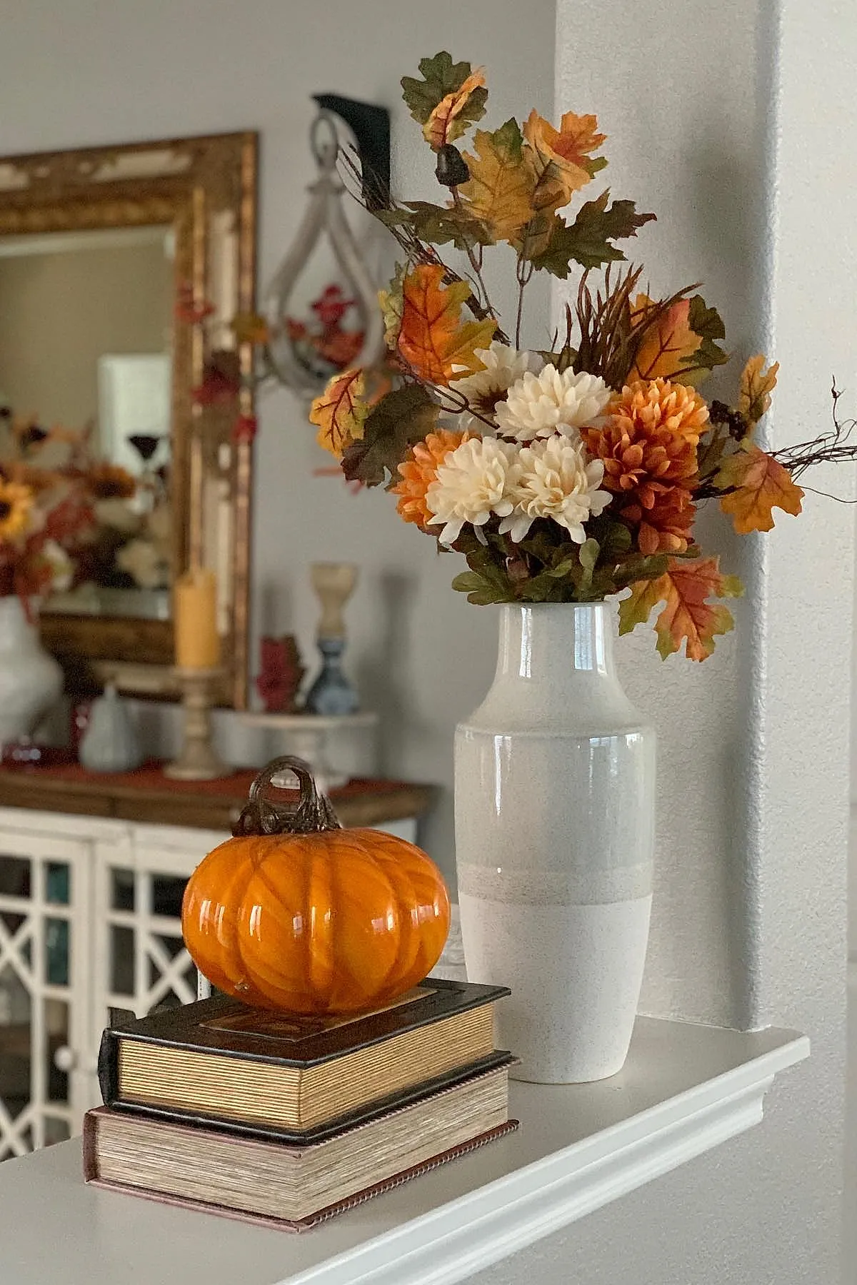 Glossy orange glass pumpkin placed on two stacked hardcover books next to a tall white ceramic vase filled with orange, cream, and green autumn flowers and leaves, set against neutral walls with blurred background furniture.