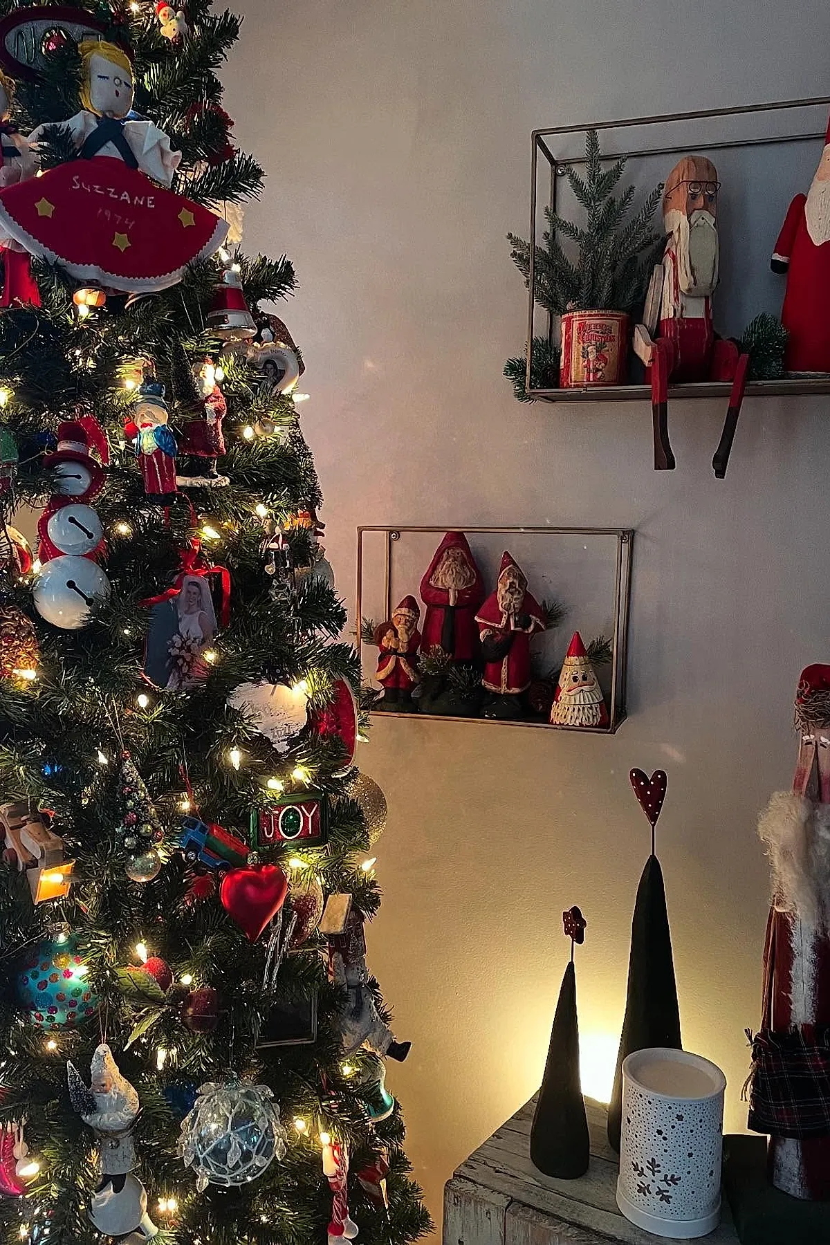 Christmas tree adorned with handmade fabric dolls, red hearts, and vintage ornaments beside floating shelves with carved santa figures and greenery for cozy antique christmas decorations.