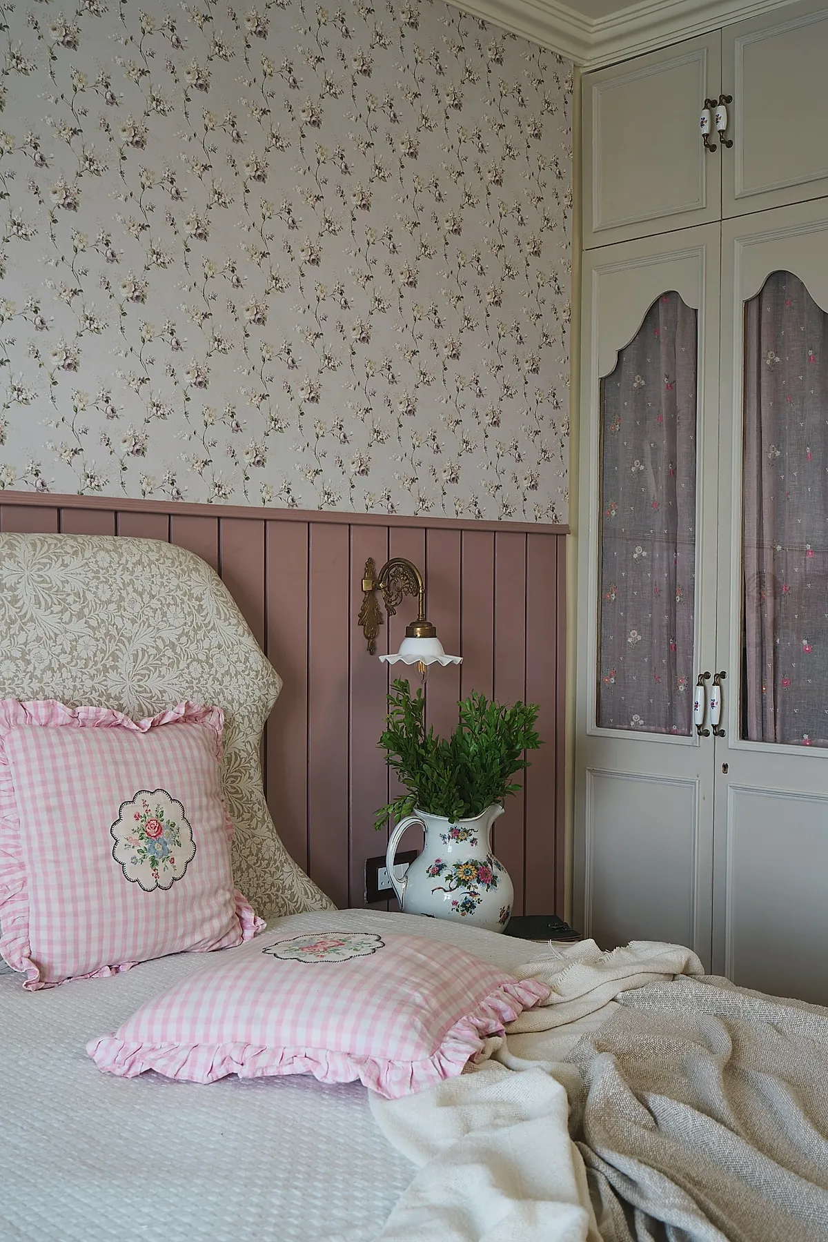 vintage bedroom corner featuring pink gingham ruffled pillows with embroidered flowers on cream quilted bed, mauve paneled wall, small brass wall lamp, floral wallpaper above paneling, white built-in wardrobe with fabric curtains and green leafy stems in ceramic pitcher