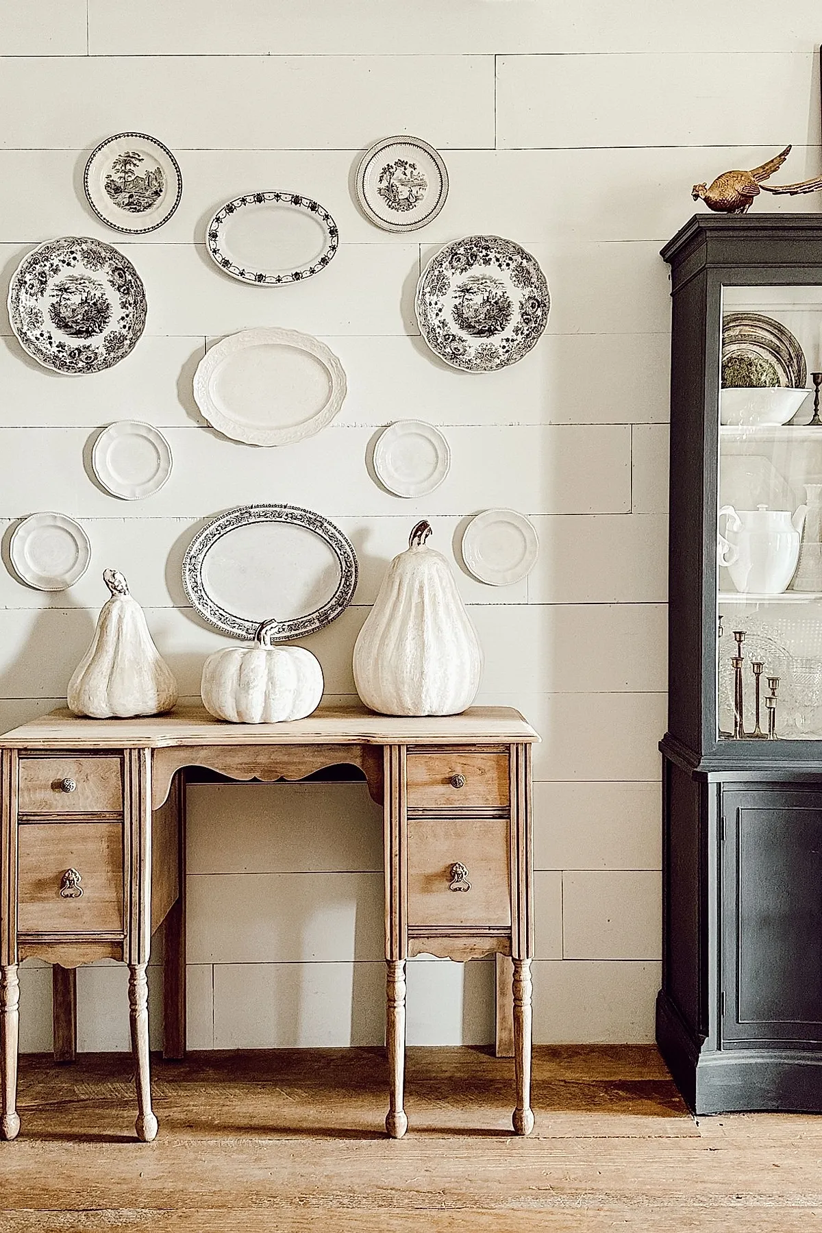 Three white pumpkins of different shapes placed on a wooden farmhouse table against a shiplap wall with black and white decorative plates arranged above.