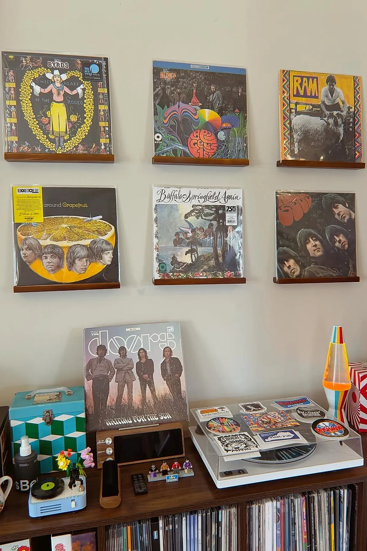 Living room wall decor ideas featuring vinyl record album covers displayed on wooden shelves arranged in two rows on a beige wall above a wooden console holding a white turntable, colorful lava lamp, blue patterned box, small figurines, and an audio device.