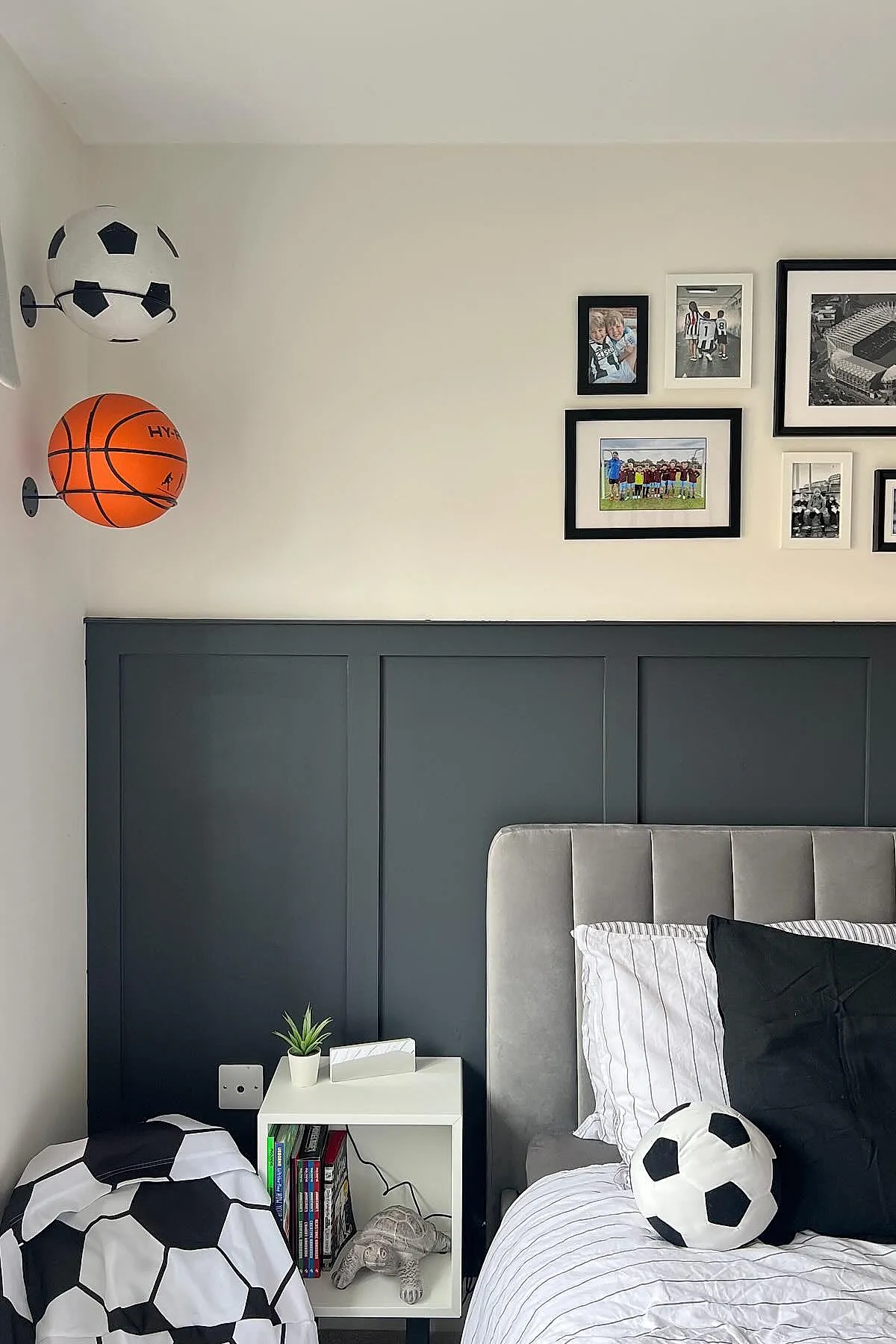 Teen boy bedroom corner featuring two sports balls mounted on the wall—a basketball and a soccer ball—above a white nightstand with books and a small plant, gray upholstered bed with striped bedding, and framed sports photos on the wall.