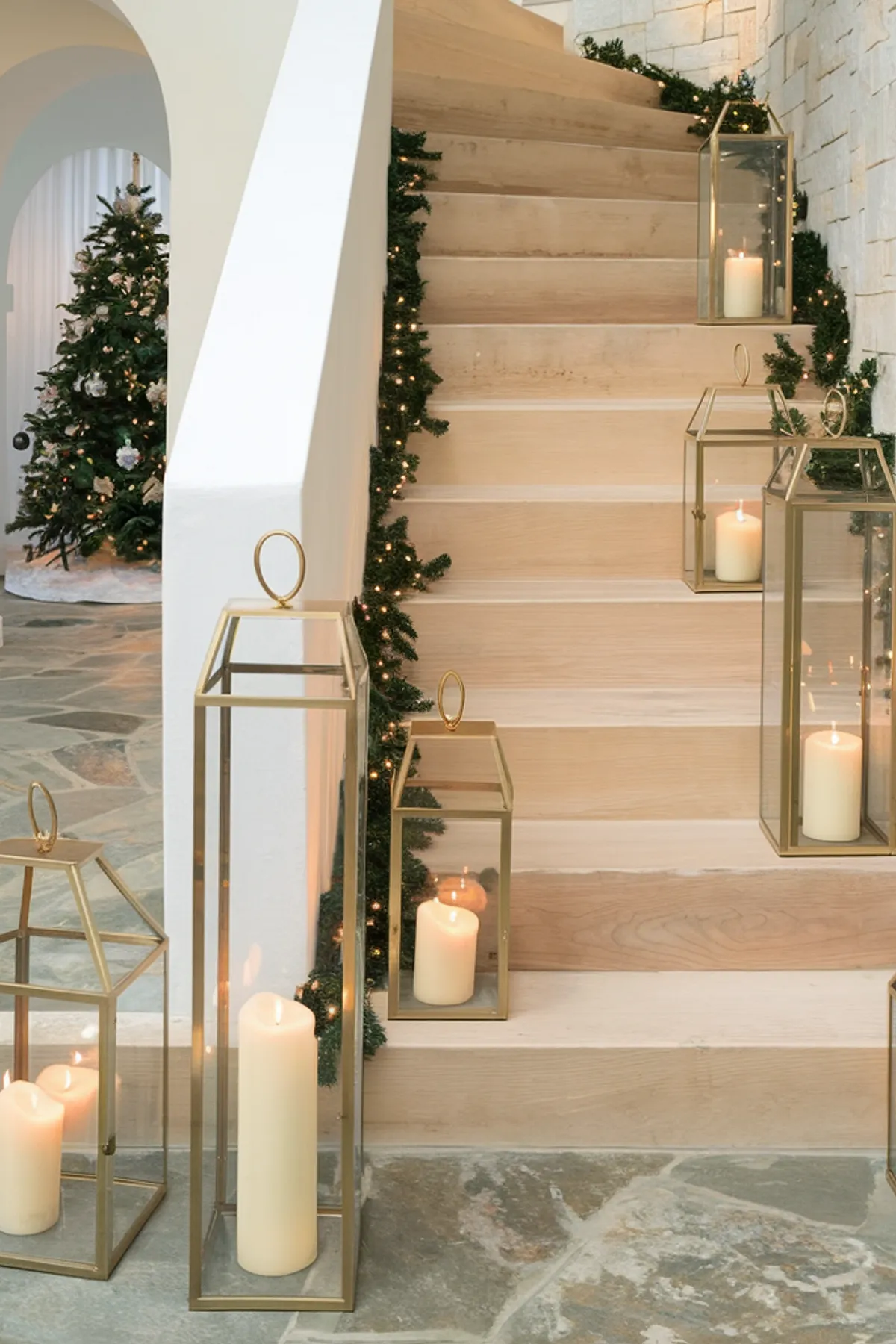 An elegant interior featuring wooden stairs adorned with golden lanterns of varying sizes, each holding lit candles that create a warm glow. The stairs are accented with garlands of greenery and fairy lights. To the left, a white archway leads to another room showcasing a decorated Christmas tree. The slate tile floor and textured walls in white and stone add to the sophisticated ambiance of the space.