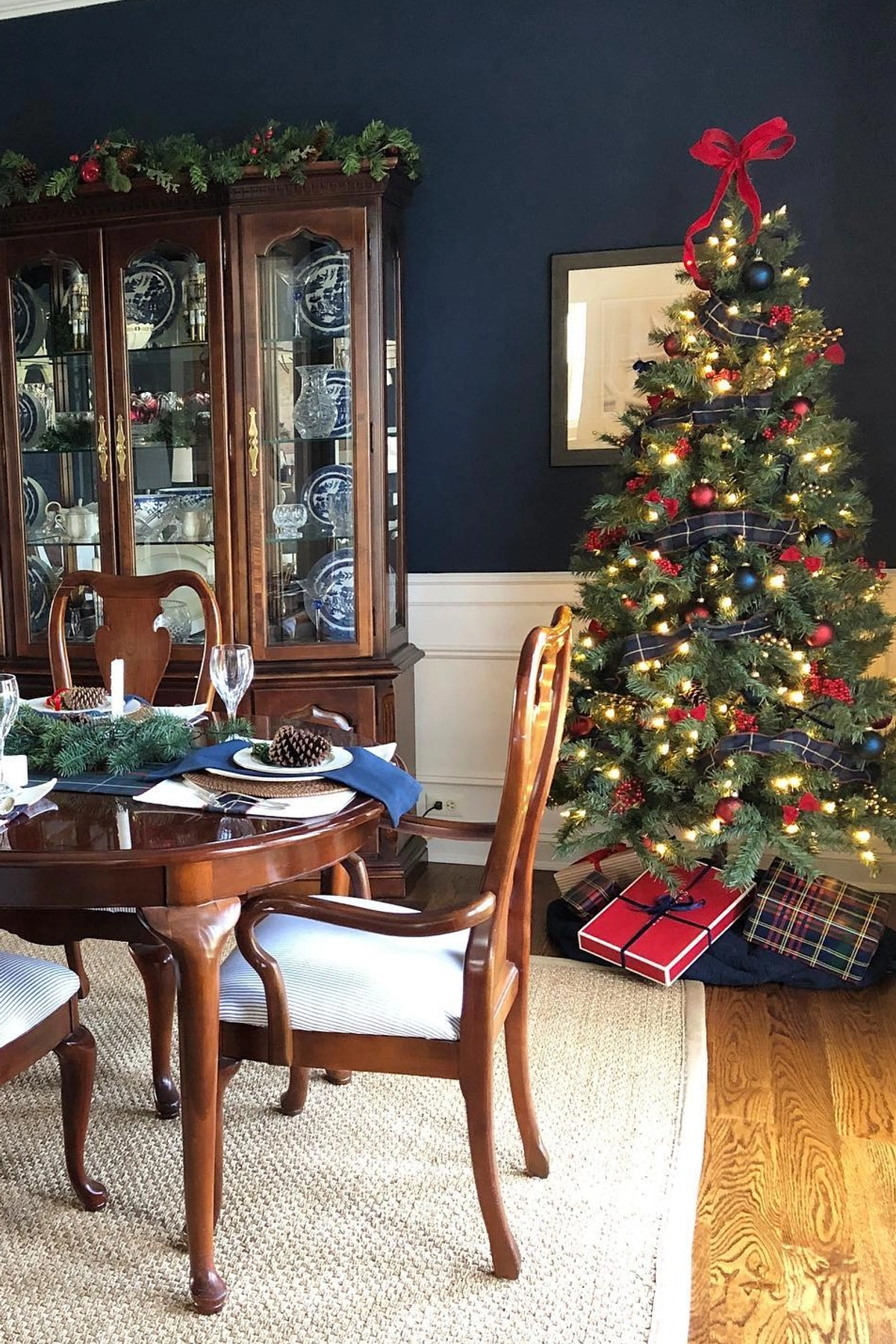 traditional christmas tree decorated with bright red berry clusters, warm white string lights, and classic round ornaments, positioned next to a dining table set with pinecones on plates and blue napkins in a navy blue walled room